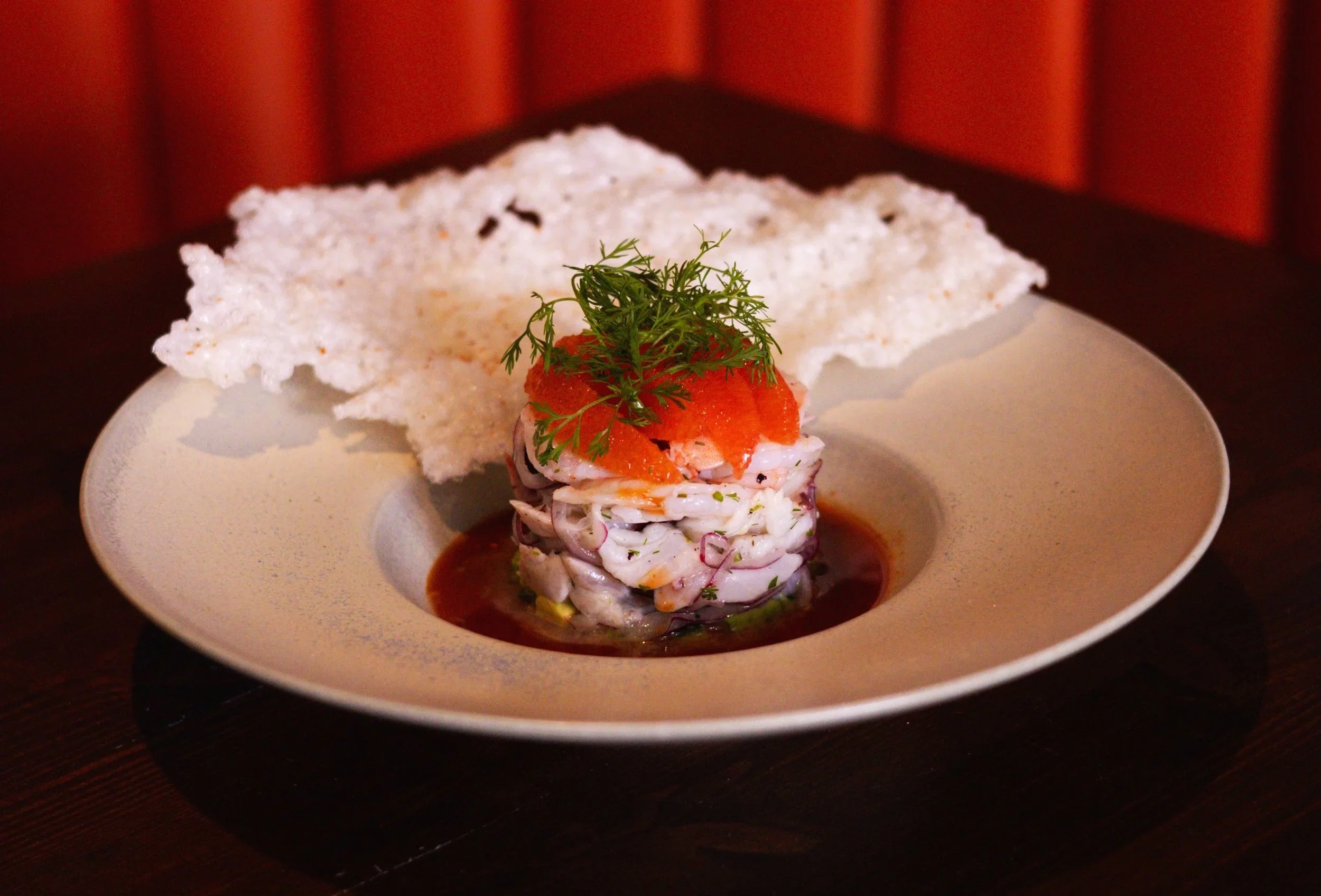 Classic Aguachile dish featuring a layered seafood salad garnished with herbs, served with a large rice paper cracker on a beige plate.