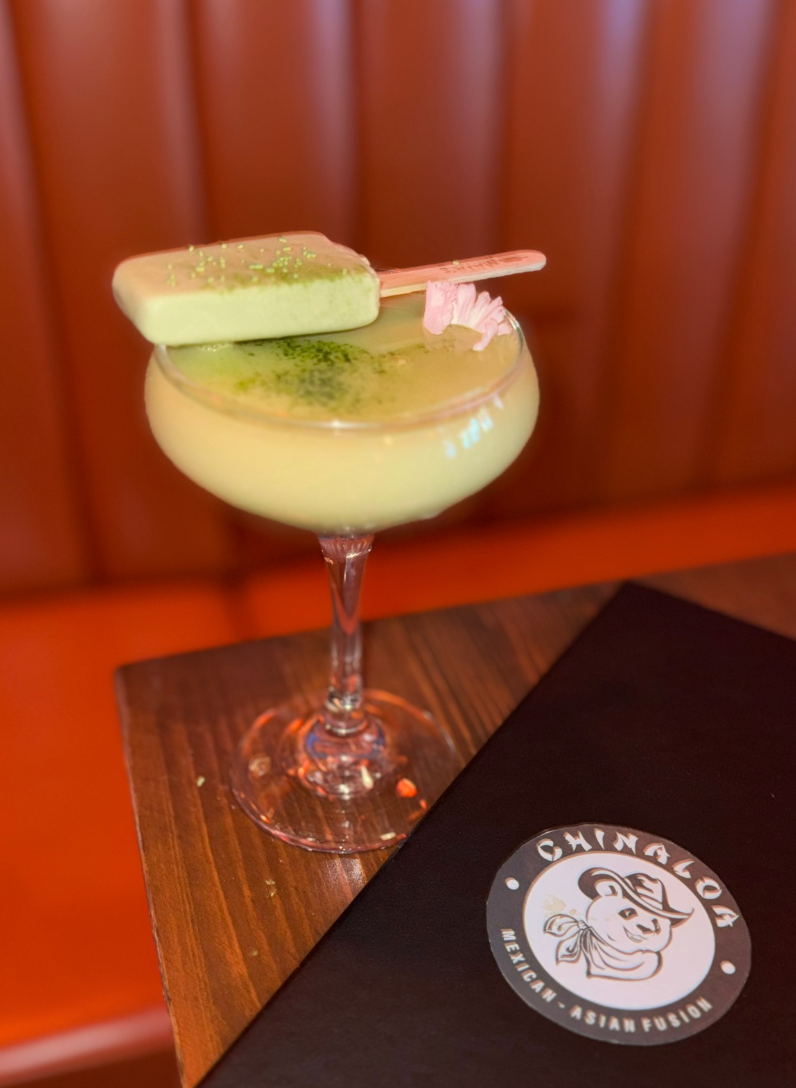 Margarita cocktail with green coloring, a popsicle garnish, on a wooden table with a black menu displaying a pig logo for Chinaglo Mexican-Asian fusion restaurant.