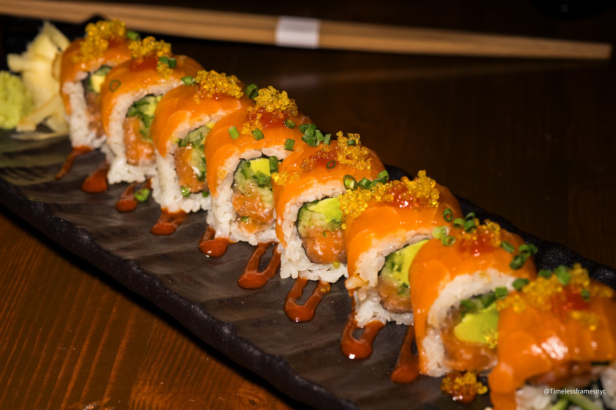 Sushi roll with salmon, avocado, and rice on a black rectangular plate, topped with orange fish roe, chopped green onions, and drizzled with sauce.