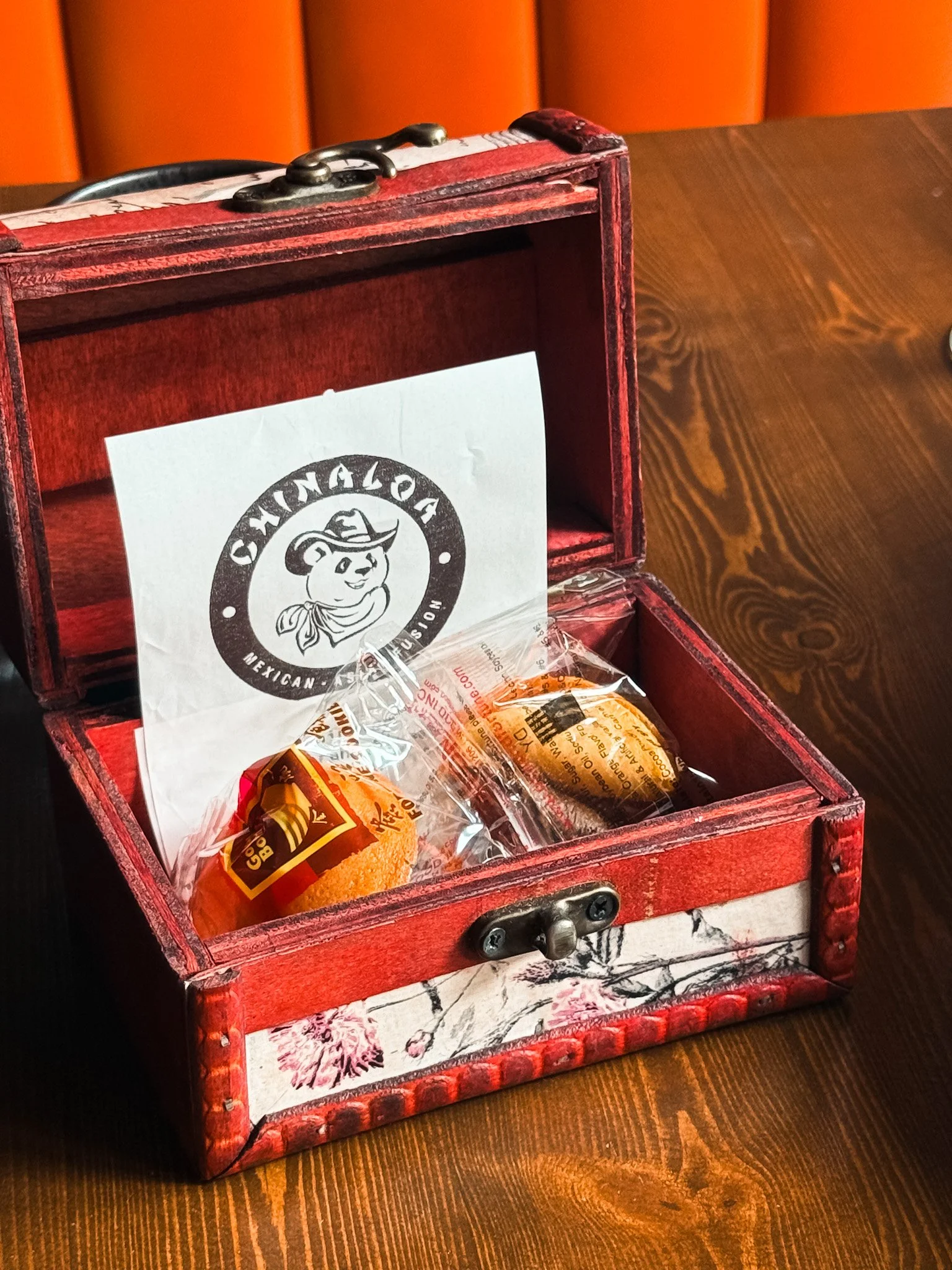 A wooden box with a red finish, decorated with floral patterns, contains two wrapped baked good items and a piece of white paper with a cartoon character wearing a cowboy hat and bandana, and text that says 'CAMPALOA MEXICAN.' The box is on a wooden 