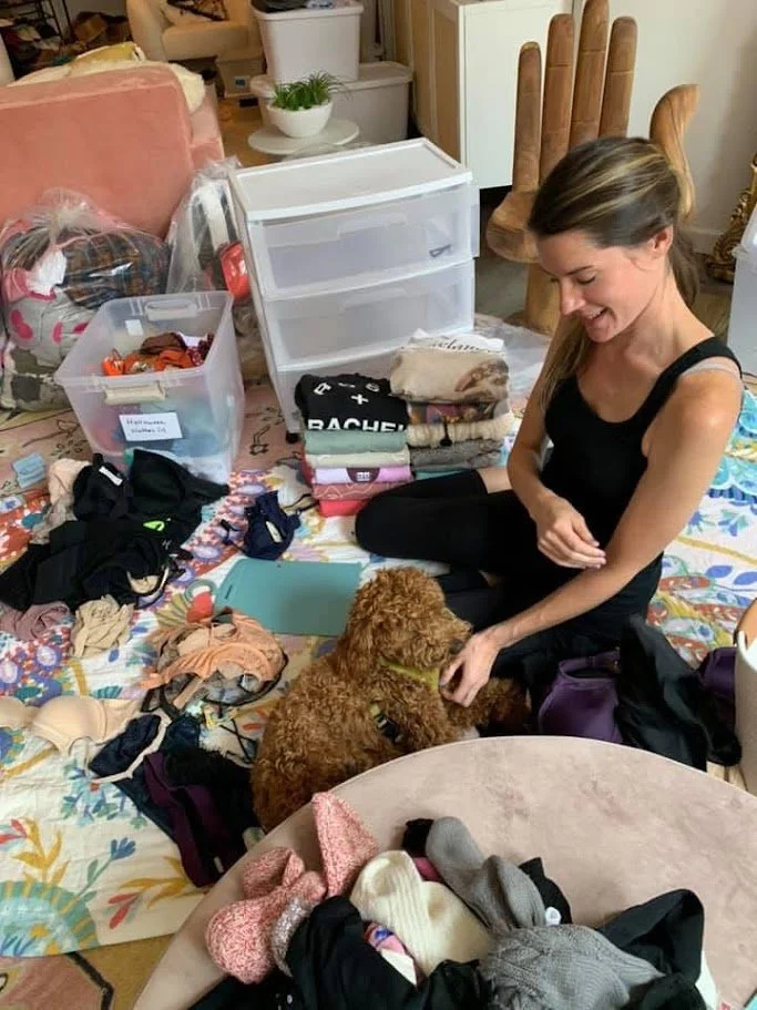 Woman sitting on the floor surrounded by clothes and belongings, playing with a small brown dog amid scattered laundry and storage containers in a cluttered room.