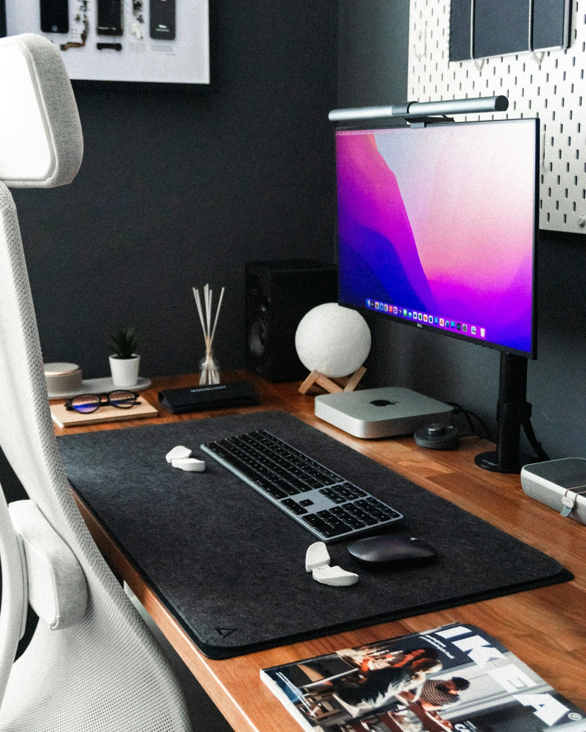 A tidy desk setup with a computer monitor, wireless keyboard and mouse, a small potted plant, glasses on a tray, a reed diffuser, and a spherical lamp on a wooden desk.