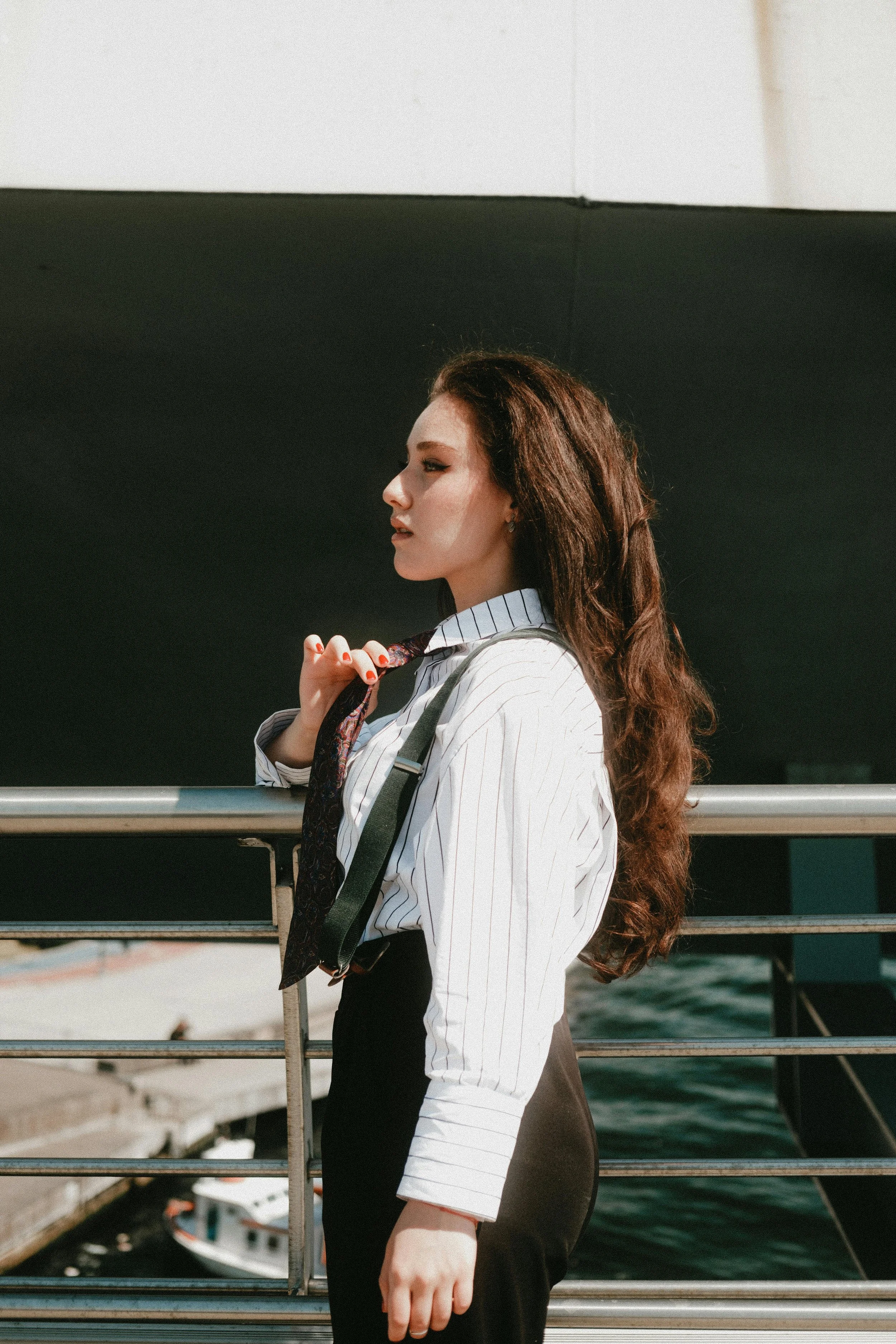 A woman with long wavy hair standing sideways on a boat or ferry, wearing a striped shirt, black skirt, and a tie, holding her tie with one hand, overlooking the water with a boat in the background.