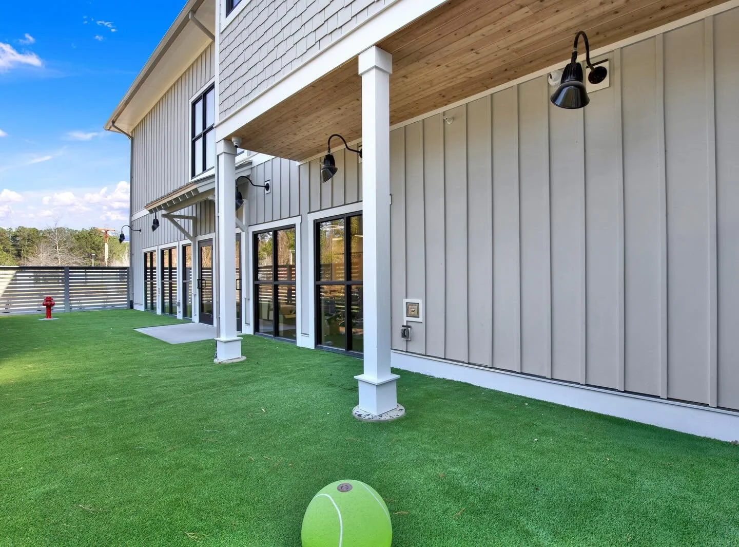 Backyard of a modern multi-story building with sliding glass doors, black outdoor wall lights, a small patio, and artificial grass. There is a red fire hydrant fountain and a tennis ball fountain on the grass of A Place To Paws boarding