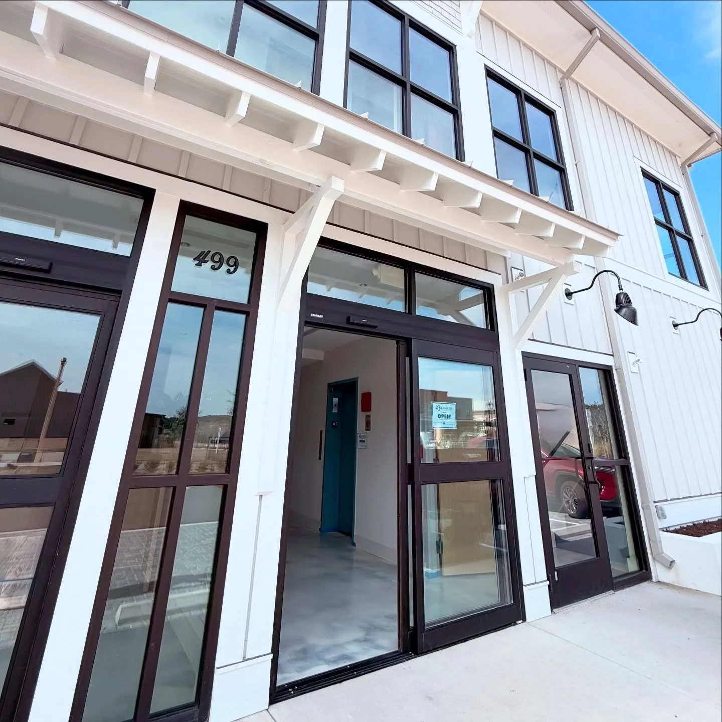 Exterior view of a modern two-story commercial building with white siding, black-framed windows, and glass doors. The building has the number 499 Buckwalter Pl Blvd near the entrance and a sign indicating it is open inside. Bluffton SC