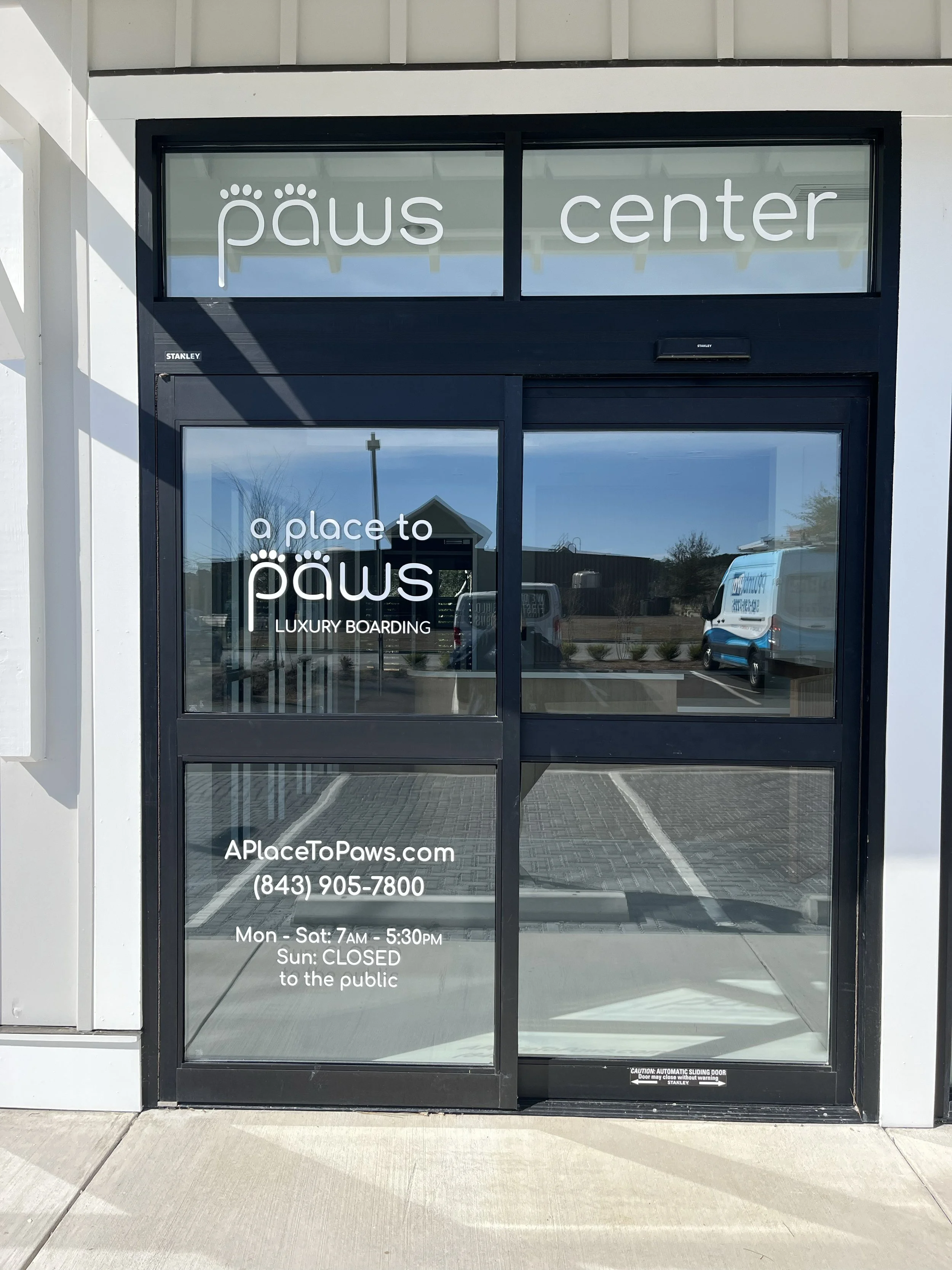 Glass door with 'paws center' on the window above, and details about the business, including the slogan 'a place to paws,' a website, phone number, and hours of operation, with reflections of parked vehicles and a building outside in Bluffton SC.