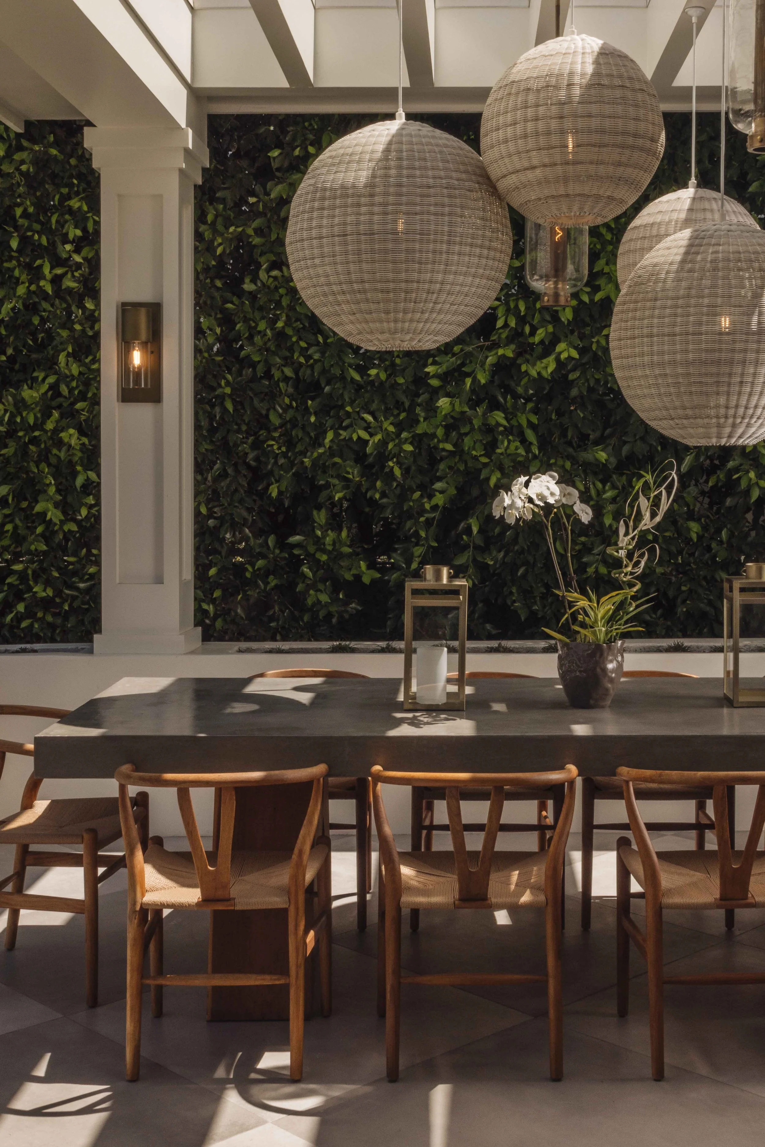 A dining table with seven wooden chairs, a potted orchid plant, and lanterns on the table, set against a backdrop of a lush green hedge, with large wicker pendant lights hanging above in an outdoor or semi-outdoor space.