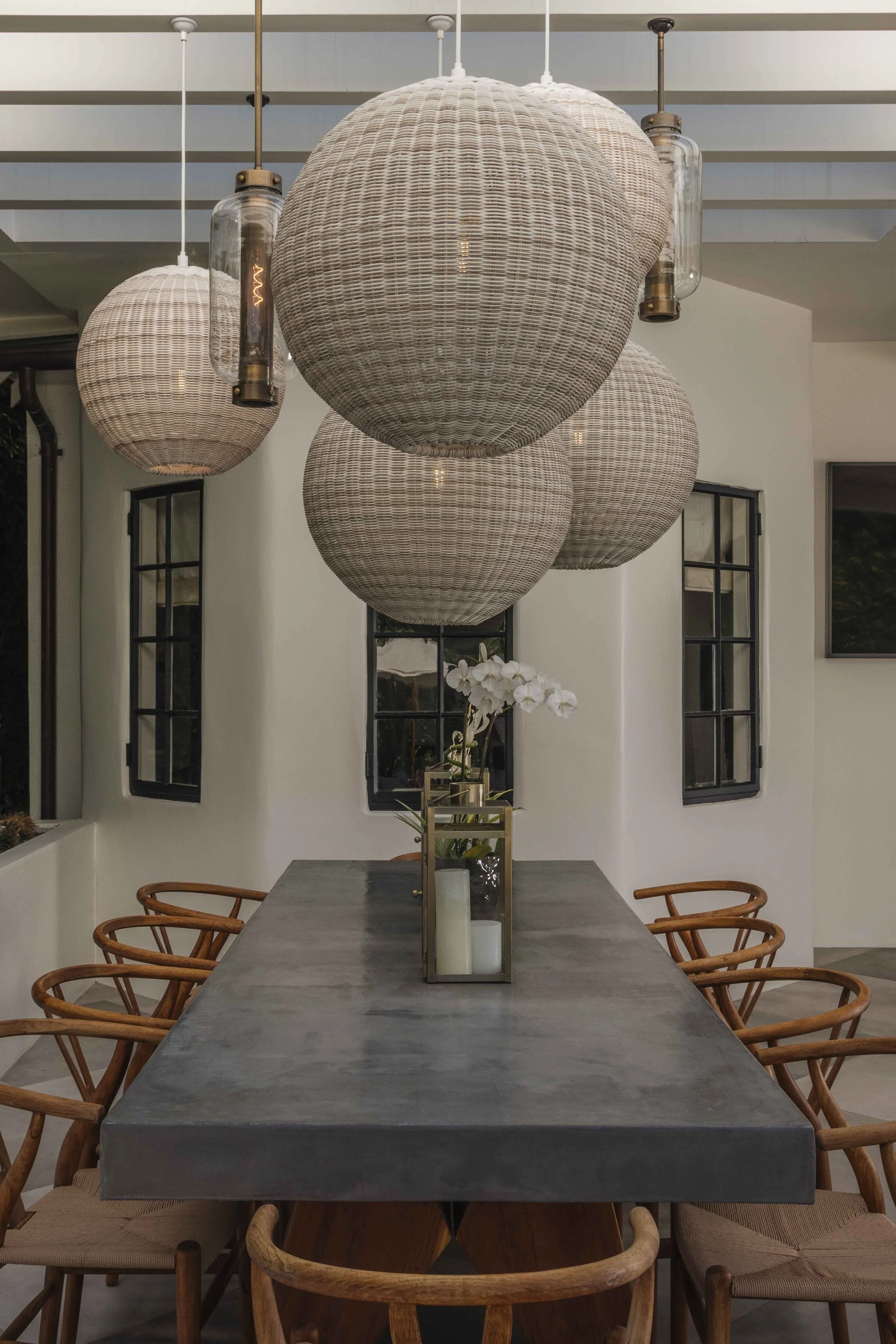 Interior dining room with a long gray table, around which are wooden chairs with woven seats, and a centerpiece of white orchids and candles. Hanging above are large woven pendant lights and two smaller glass and metal fixtures.