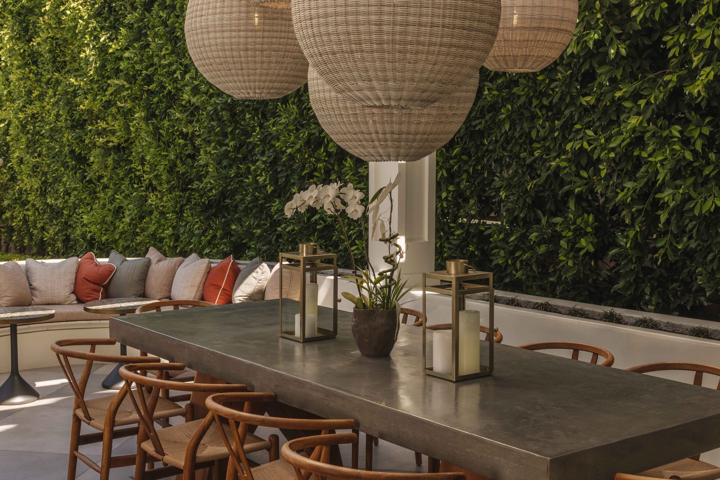 Outdoor patio with a long table, surrounded by wooden chairs, decorated with a potted orchid and lanterns, with a cushioned bench seating area with pillows, and hanging woven pendant lights above, against a green hedge background.