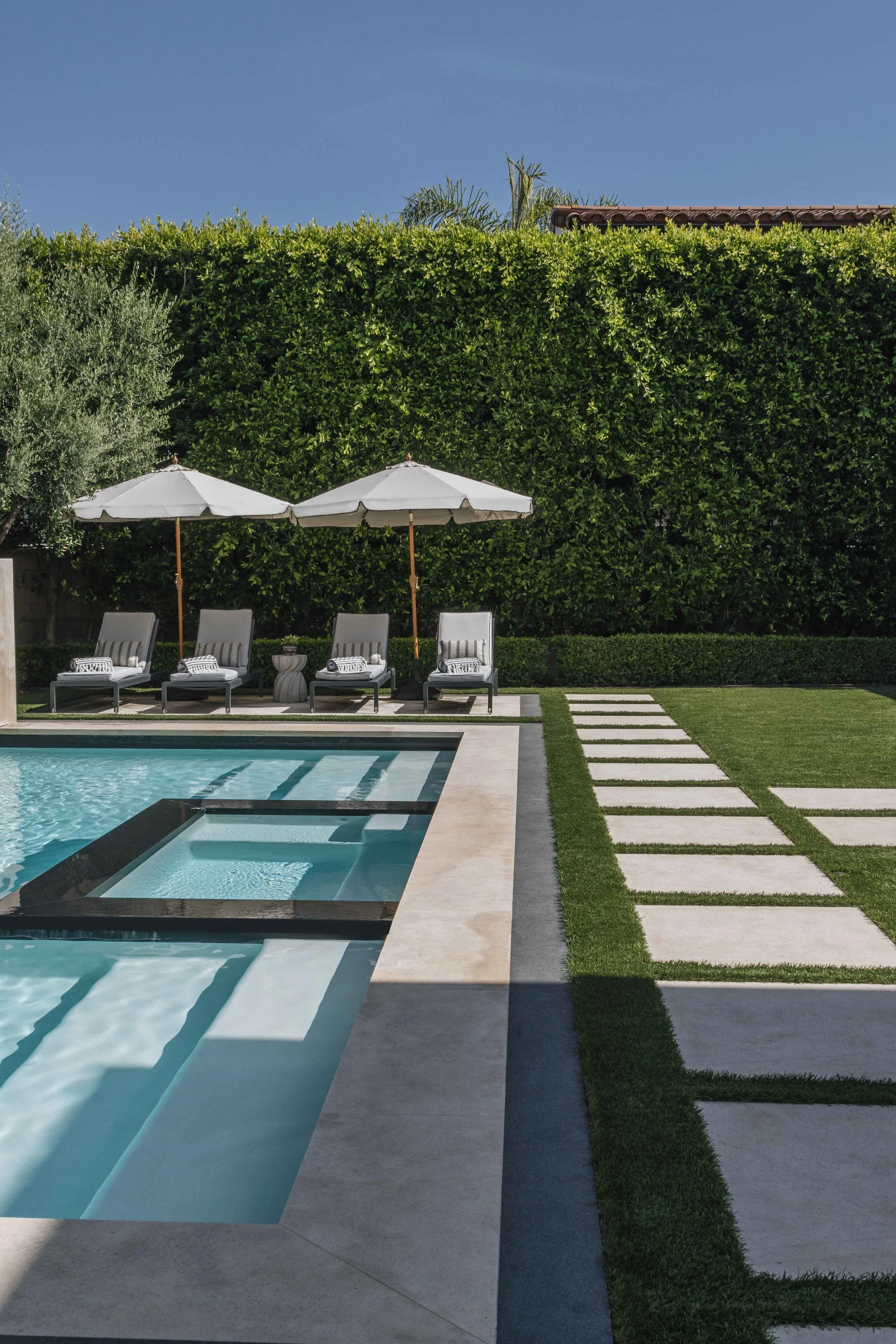 A backyard swimming pool area with lounge chairs and umbrellas, surrounded by lush green hedges and a well-manicured lawn.