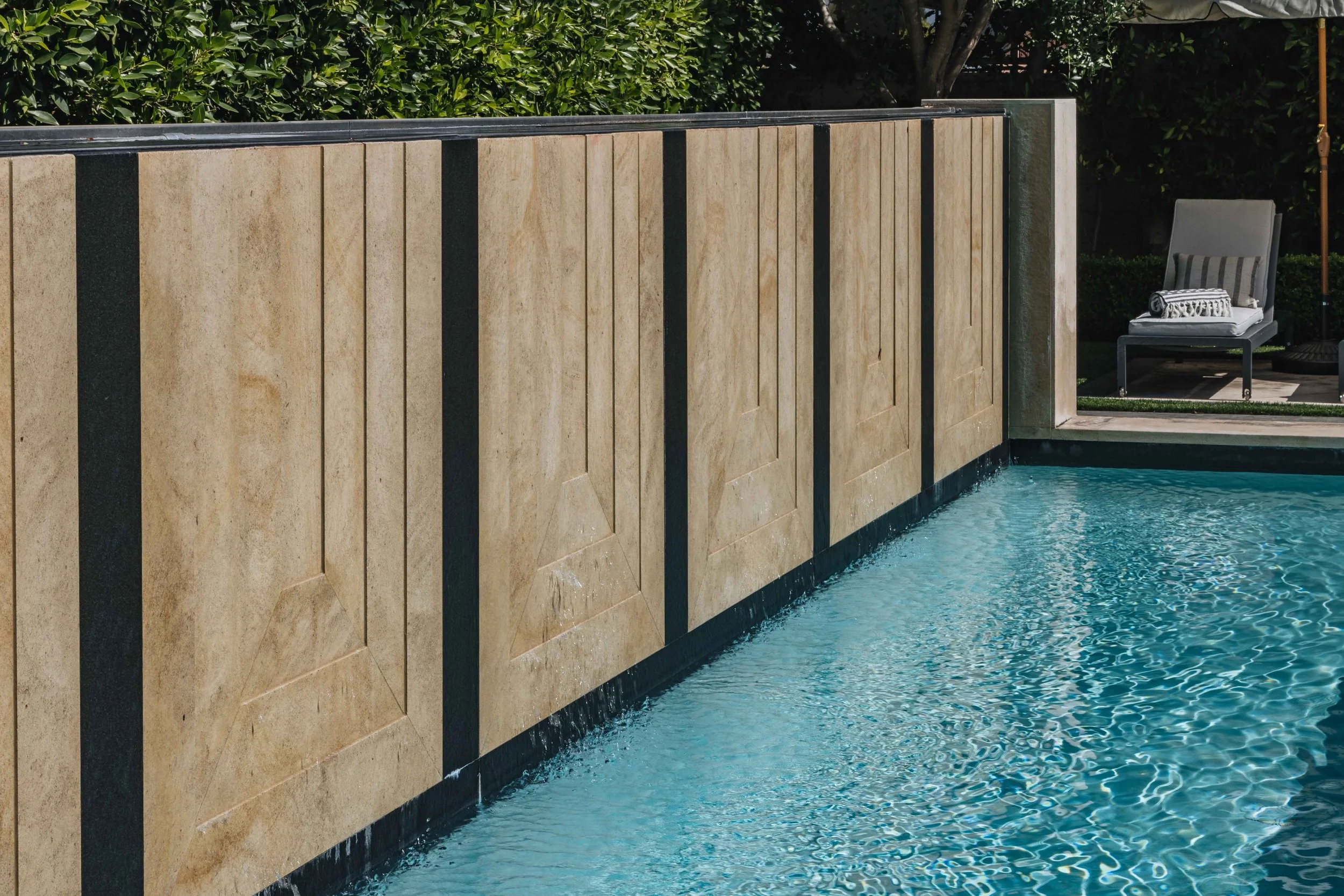 Close-up of a swimming pool with a beige stone wall and a lounge chair in the background.