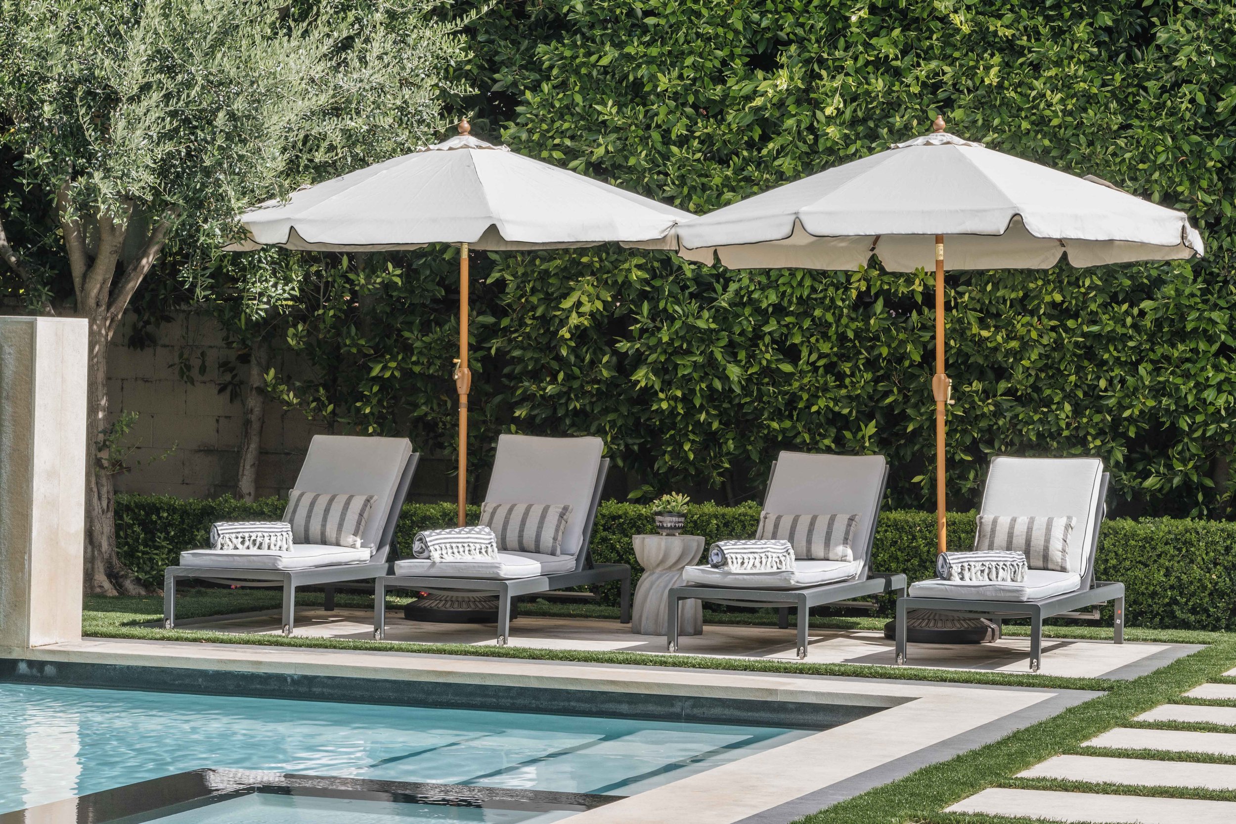 Four lounge chairs with striped pillows and rolled towels, shaded by two large white umbrellas, next to a swimming pool with a lush green hedge and trees in the background.