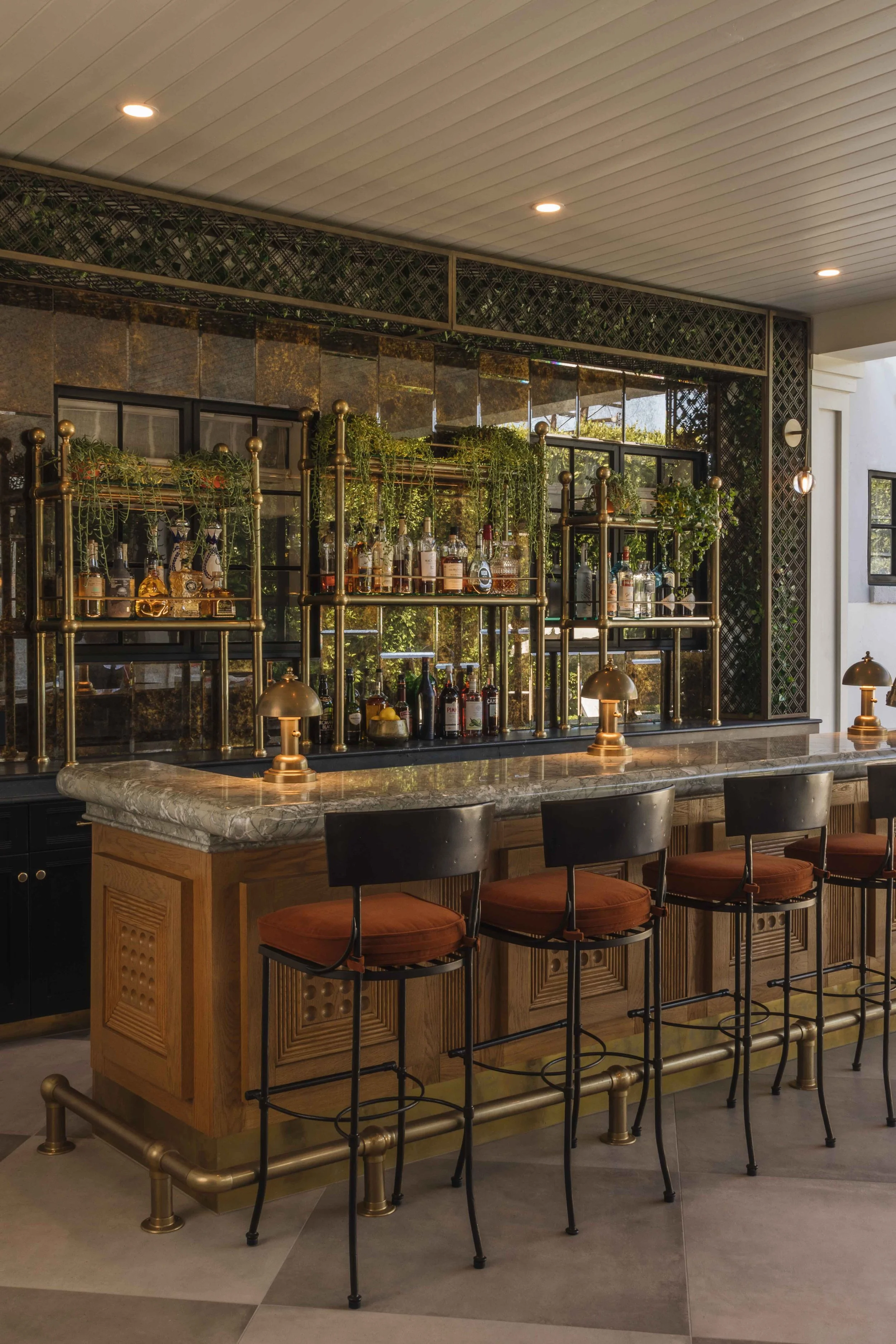 A stylish bar with a marble countertop, black chairs with red cushions, and a gold metal and glass shelving unit behind it displaying bottles of alcohol and decorative plants.