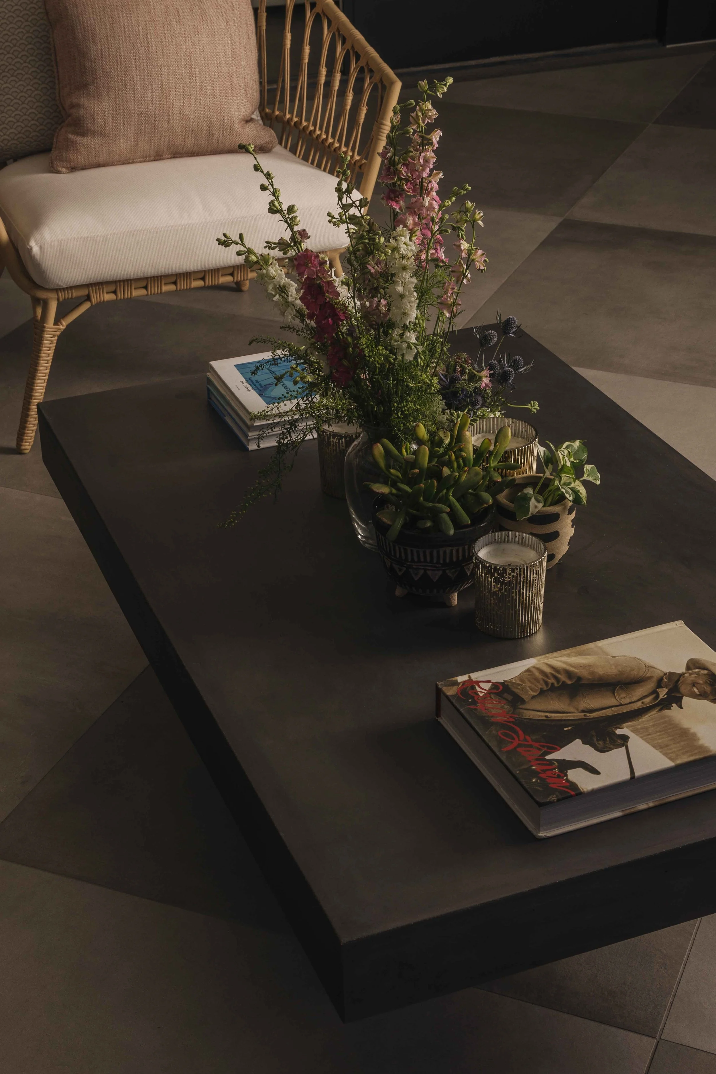 A dark coffee table with a floral arrangement, a magazine featuring a person holding a leash, and small decorative candle holders. A beige woven chair with pillows is in the background.