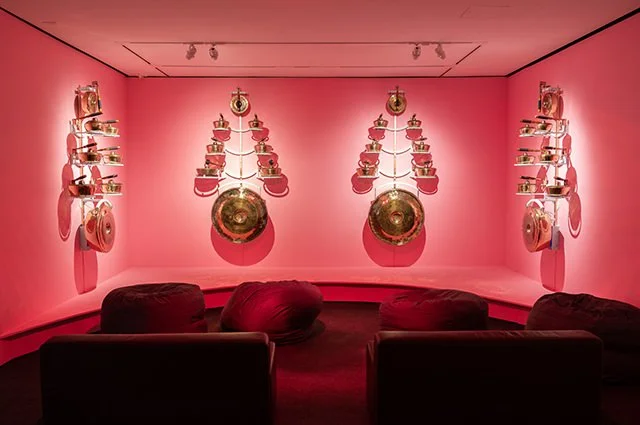 An art installation with pink walls displaying various vintage kitchenware, including pots, pans, and teapots, arranged on shelves and hanging on the walls, with seating in the foreground.