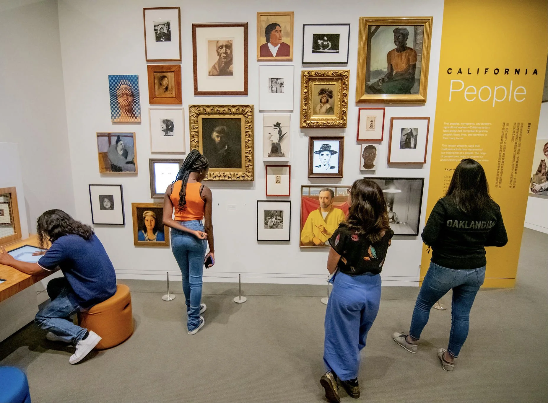 People viewing an art exhibit in a museum, featuring a wall filled with framed portraits and photographs of diverse individuals, with a yellow informational panel on the right.