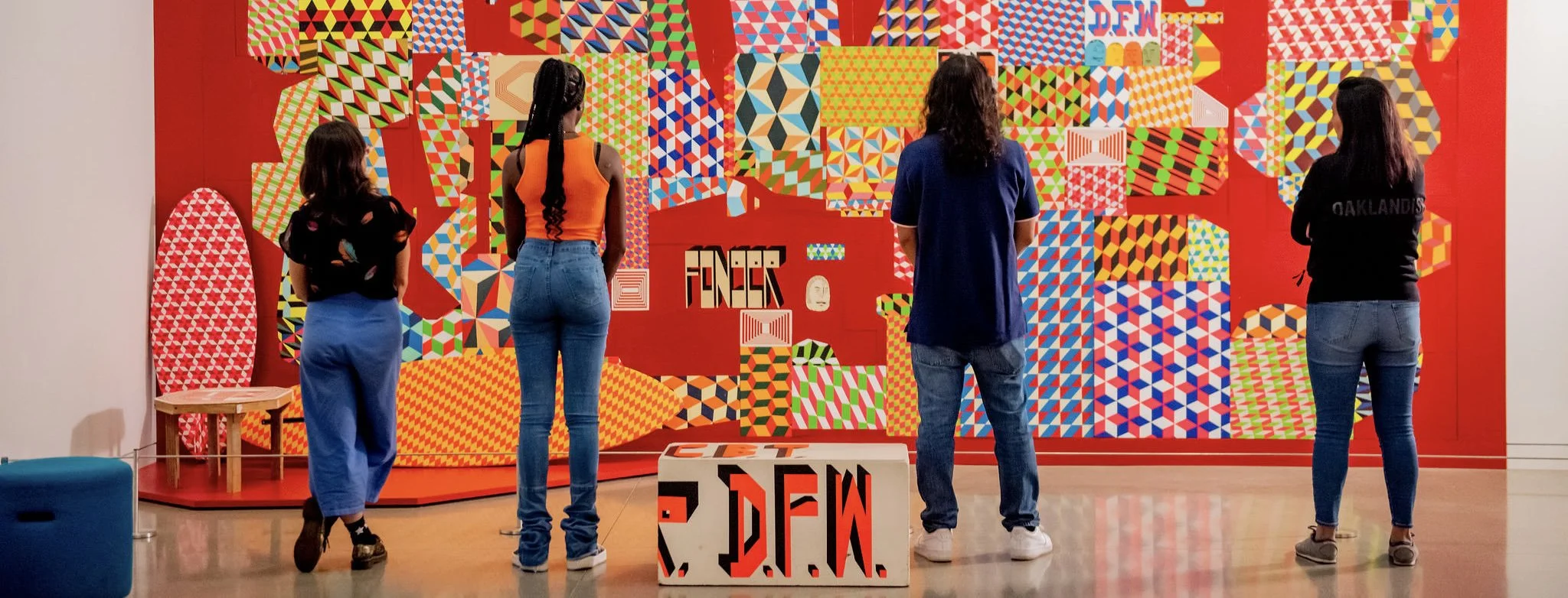 Four women stand facing a colorful abstract artwork in an exhibition space. The artwork features various geometric patterns on a red background. One woman has braids and wears an orange top and jeans, another has long dark hair wearing a black top an