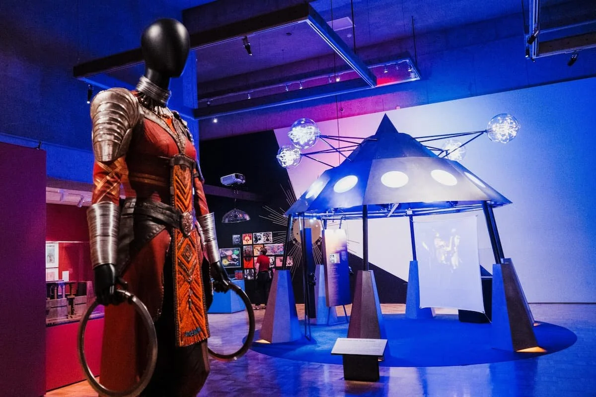 A display in a museum showing a suit of armor and a spaceship model with glowing lights and hanging orbs.