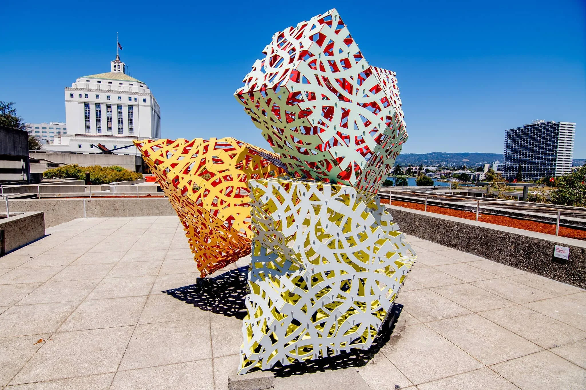 Colorful abstract metal sculptures with intricate cutout patterns, one orange and the other yellow, set against a cityscape with buildings and a clear blue sky.