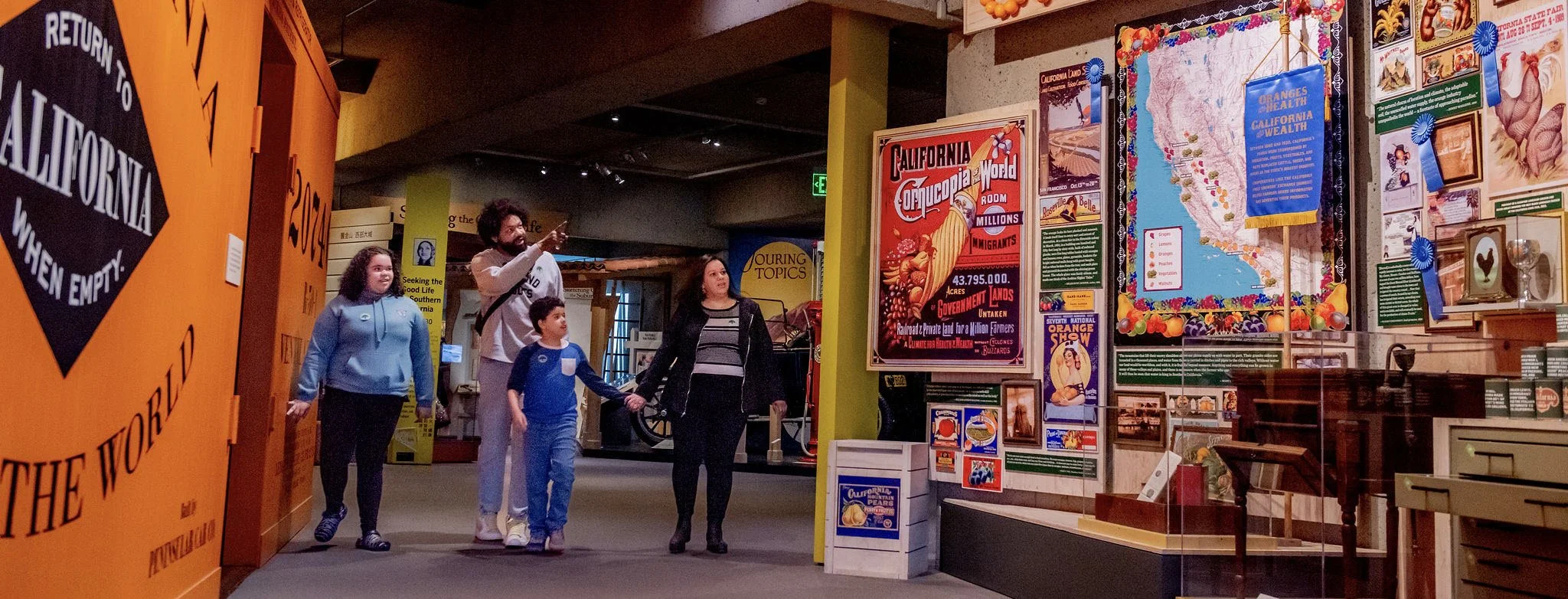 A group of four people, including two children and two adults, walking through a museum exhibit area with colorful posters and displays about California attractions and history.