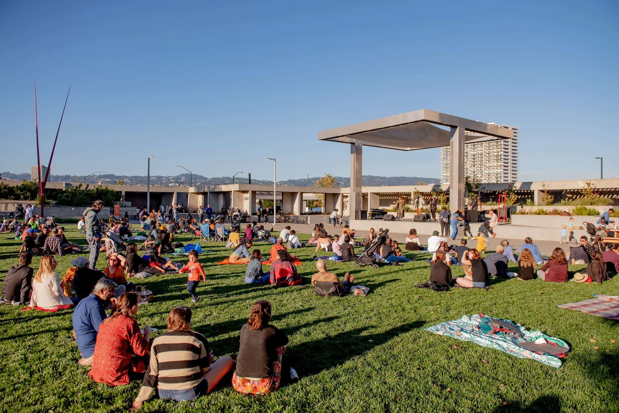 People gathered on a grassy area for an outdoor event with a stage and a modern pavilion under a clear blue sky.