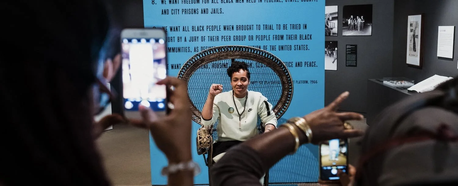 A woman with short hair and a white sweatshirt sits in an ornate chair, smiling and flexing her arm. Two people are taking photos of her with their phones in an art gallery or exhibit space, with a blue background and framed photos on the wall behind