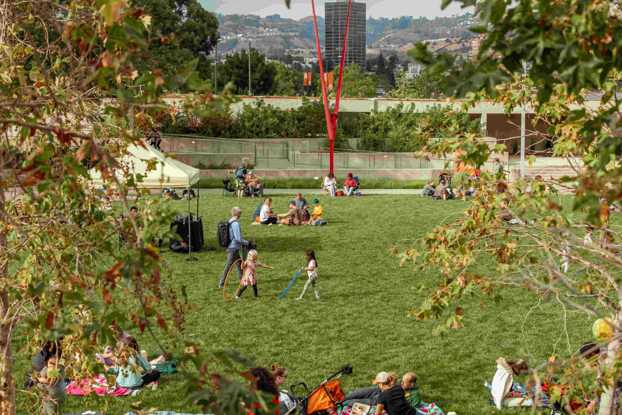 People relaxing and playing on a grassy park, some sitting on mats, children playing with hula hoops, surrounded by trees with city buildings and hills in the background.
