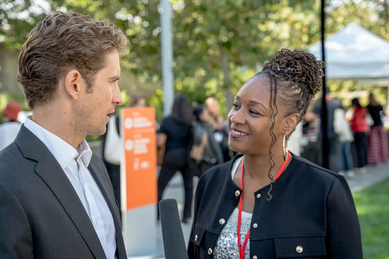 A man in a suit being interviewed by a woman at an outdoor event, with a microphone held near him and people in the background.