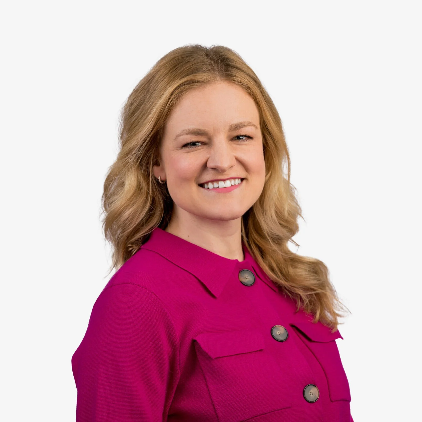 A woman with wavy, shoulder-length blonde hair, smiling, wearing a bright pink blazer with large dark buttons, against a plain white background.