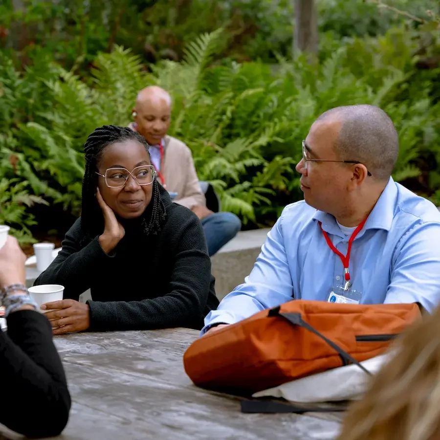 People engaging in conversation outdoors with lush green foliage in the background.