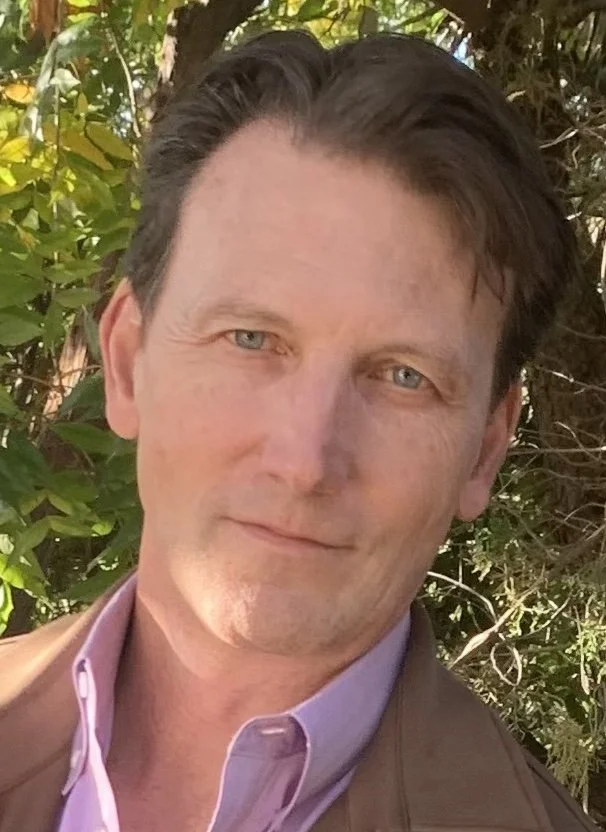 Close-up portrait of a middle-aged man with dark hair and a light beard, smiling outdoors with a blurred background.