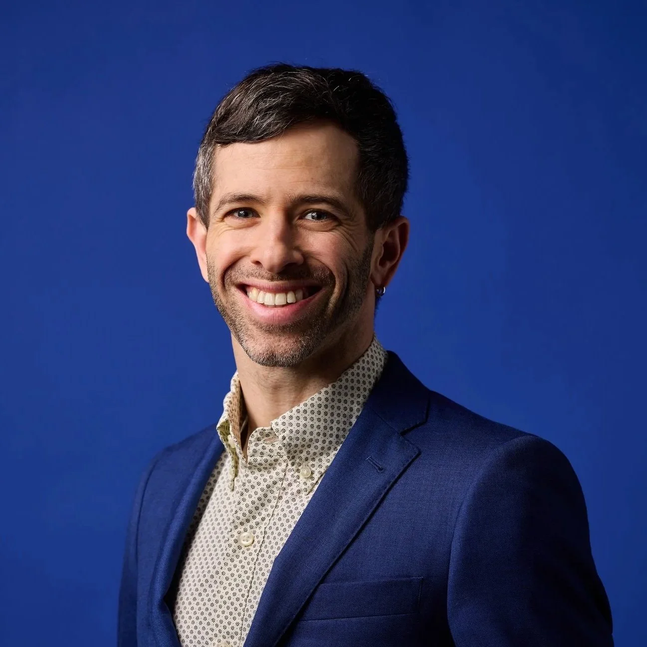 A smiling man wearing a blue jacket and backpack taking a selfie on a hiking trail surrounded by trees.