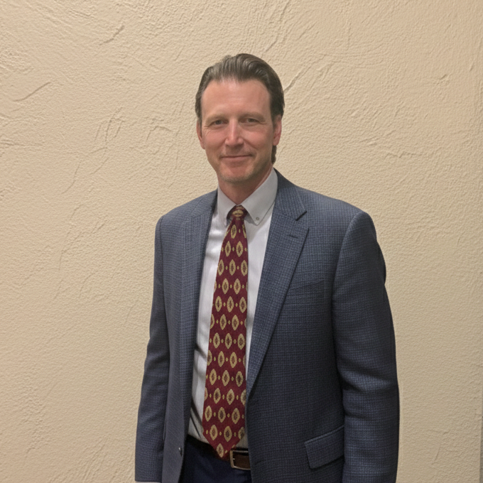 Close-up portrait of a middle-aged man with dark hair and a light beard, smiling outdoors with a blurred background.