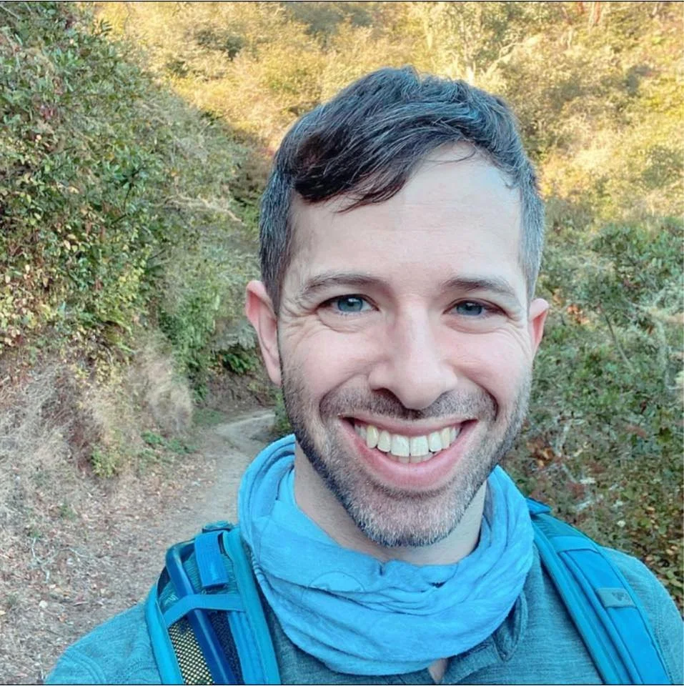 A smiling man wearing a blue jacket and backpack taking a selfie on a hiking trail surrounded by trees.
