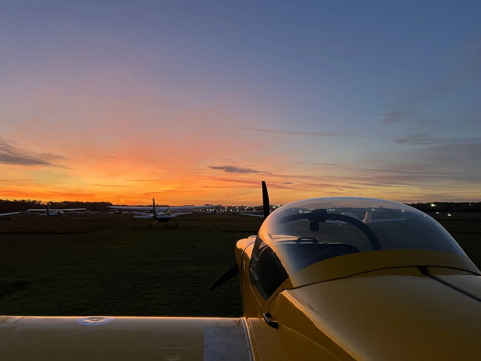 A yellow glider aircraft on a grass field at sunset, with a row of other gliders in the background under a colorful sky.