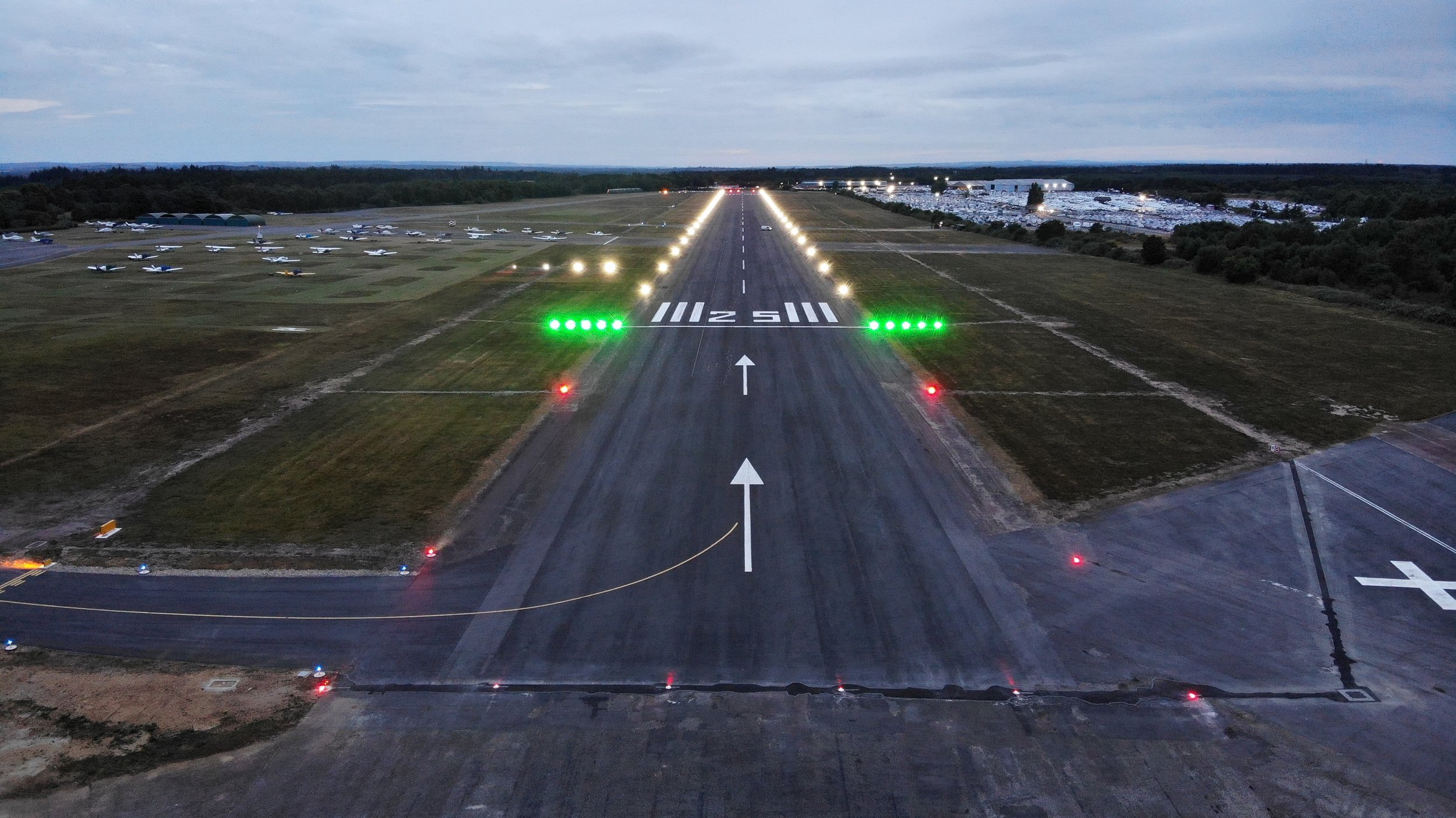 View of an airport runway with aircraft parked to the side, illuminated by runway lights, with cloudy sky overhead.