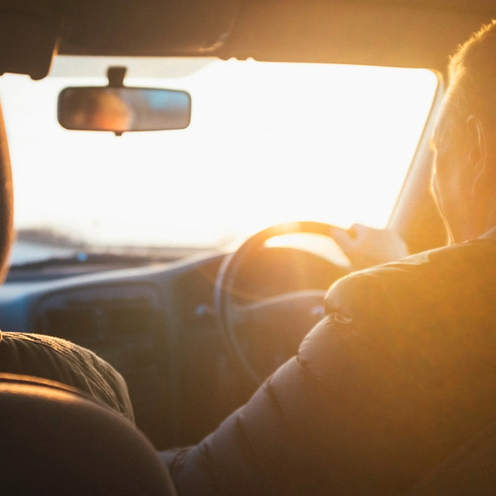 View from inside a car showing two people, one driving, during sunset.