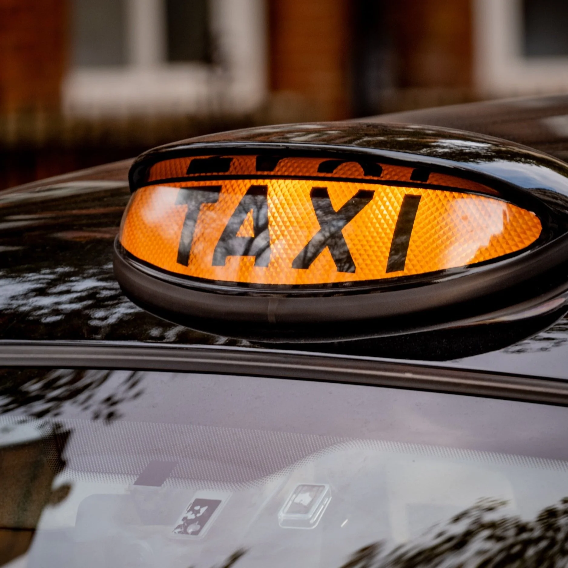 Close-up of a taxi sign on top of a black car, illuminated in orange with black letters spelling 'TAXI'.