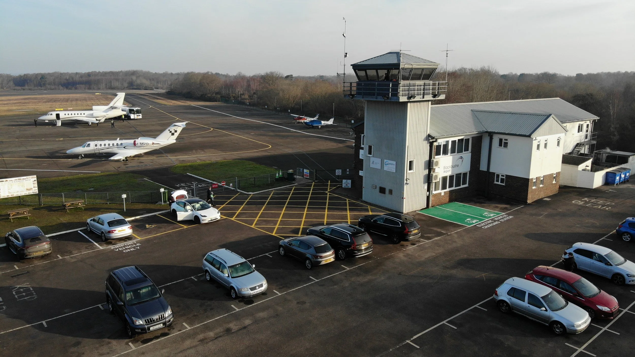 Parking lot and terminal building at a small airport with several parked cars, parked small jets, and a control tower.