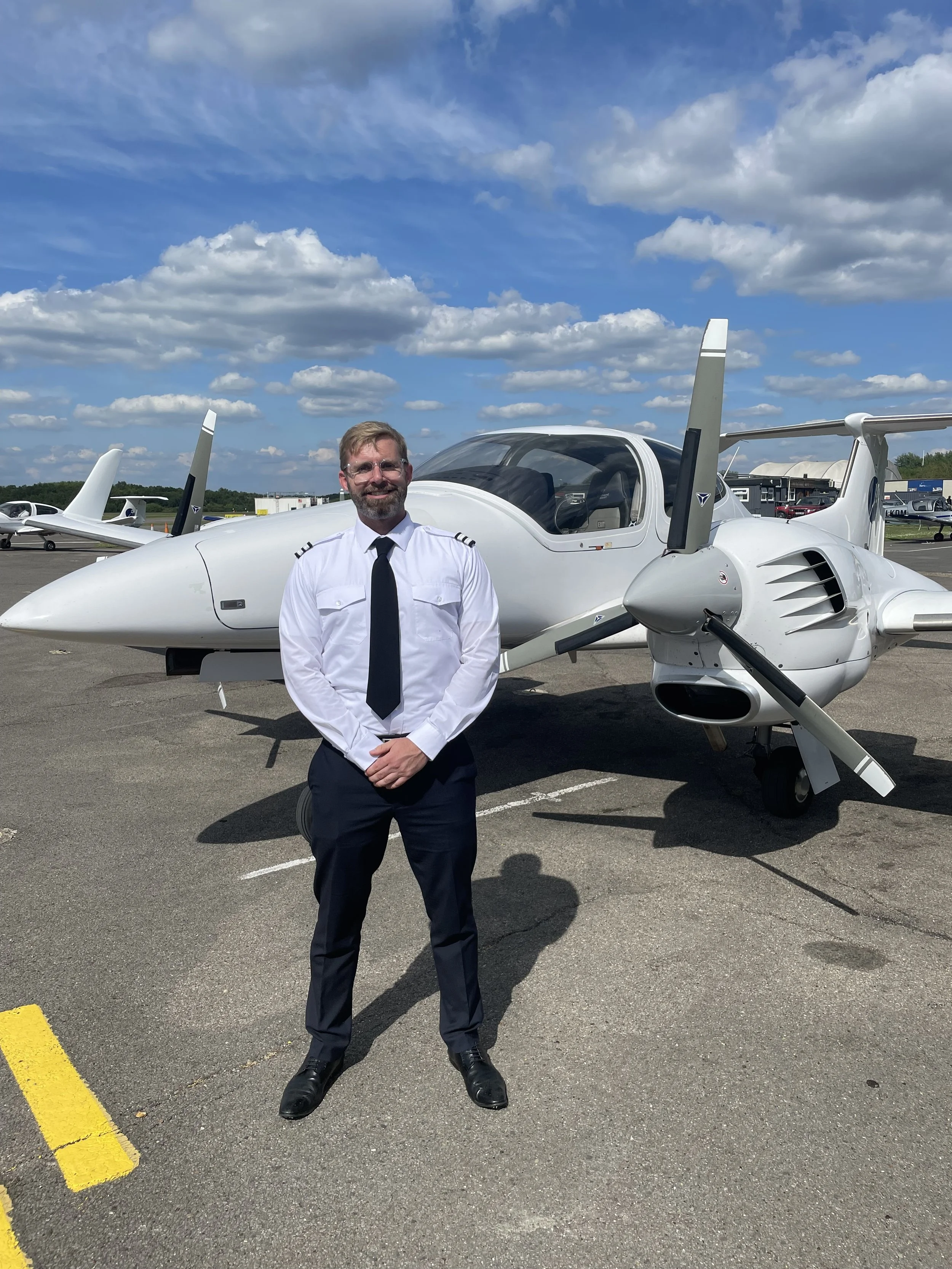 A man dressed as a pilot in uniform standing in front of a small white private jet on an airport tarmac, with other aircraft and planes visible in the background under a partly cloudy sky.