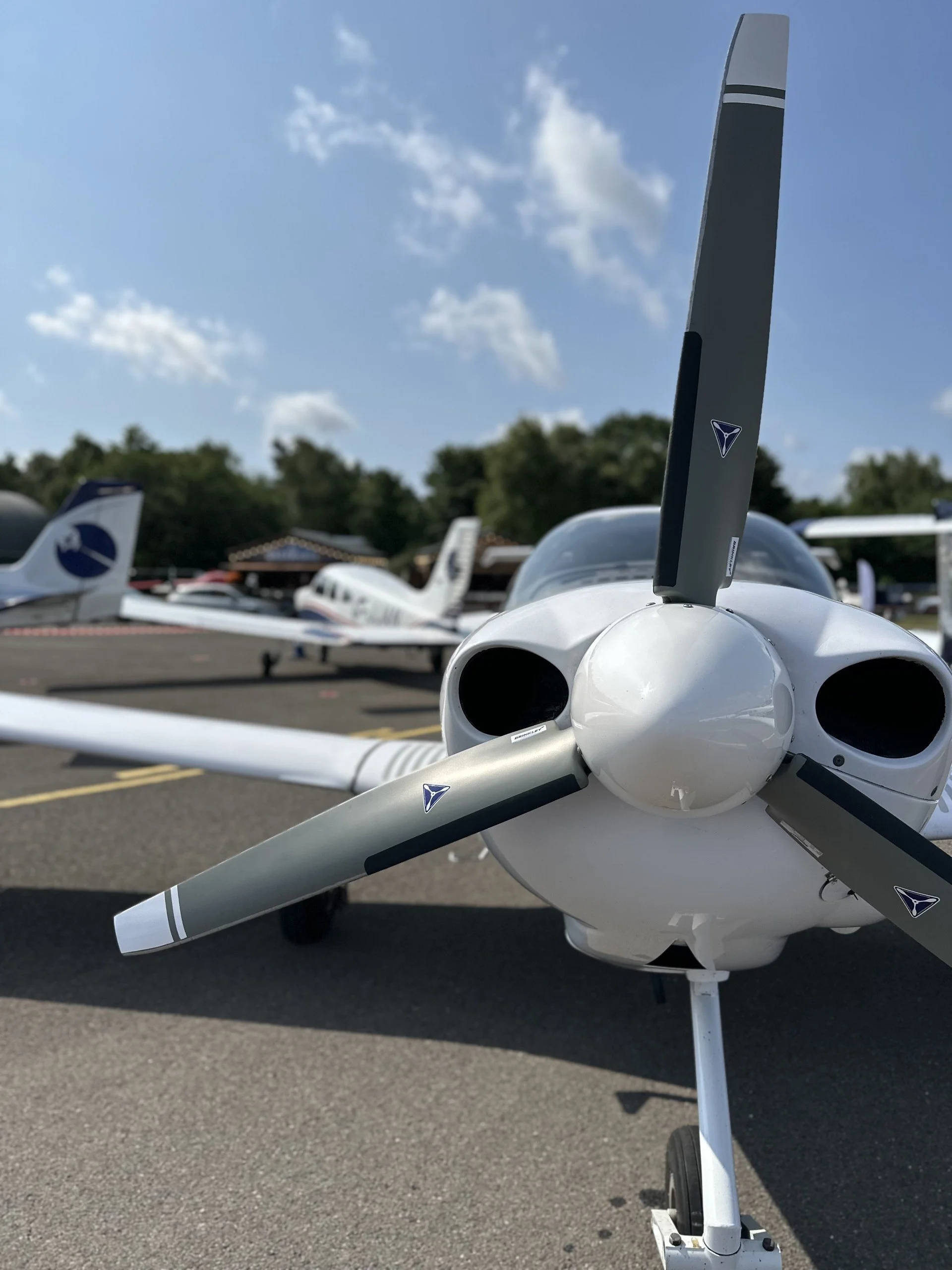 Front view of a small white airplane with a propeller, parked on tarmac at a small airport, with several other small airplanes in the background and trees under a partly cloudy sky.