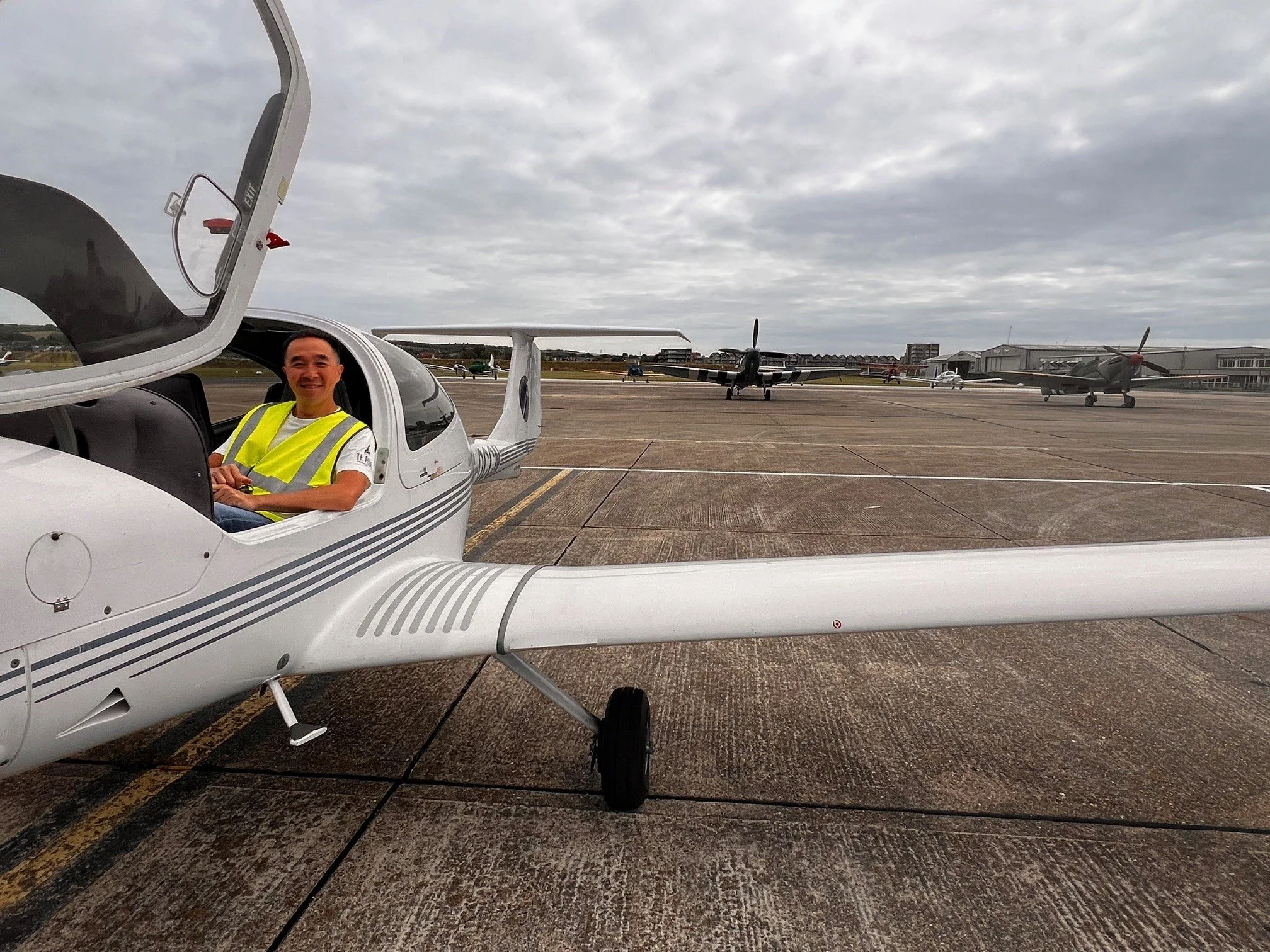 Person sitting inside a small white aircraft on the tarmac of an airport, with a gray cloudy sky and other airplanes in the background.