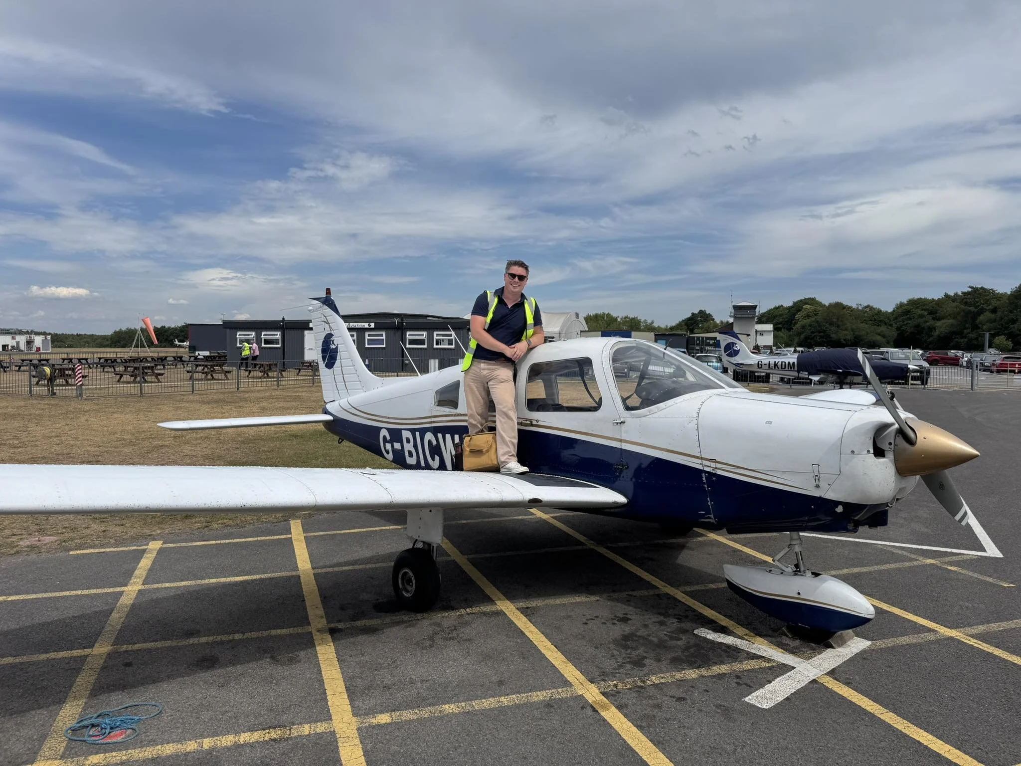 A man in a high-visibility vest standing on the wing of a small white and blue private jet at an airport, with a person walking in the background and several other small airplanes parked nearby under a partly cloudy sky.