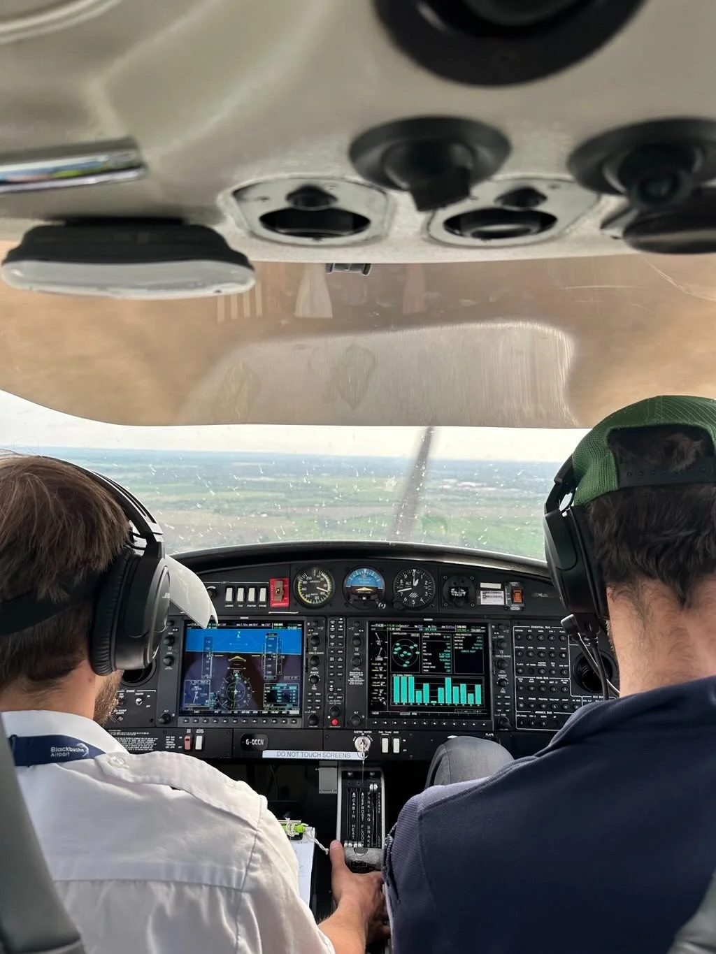 Inside the cockpit of a small airplane, with two pilots wearing headsets. The view shows the instrument panel, with screens and gauges, and the runway outside.