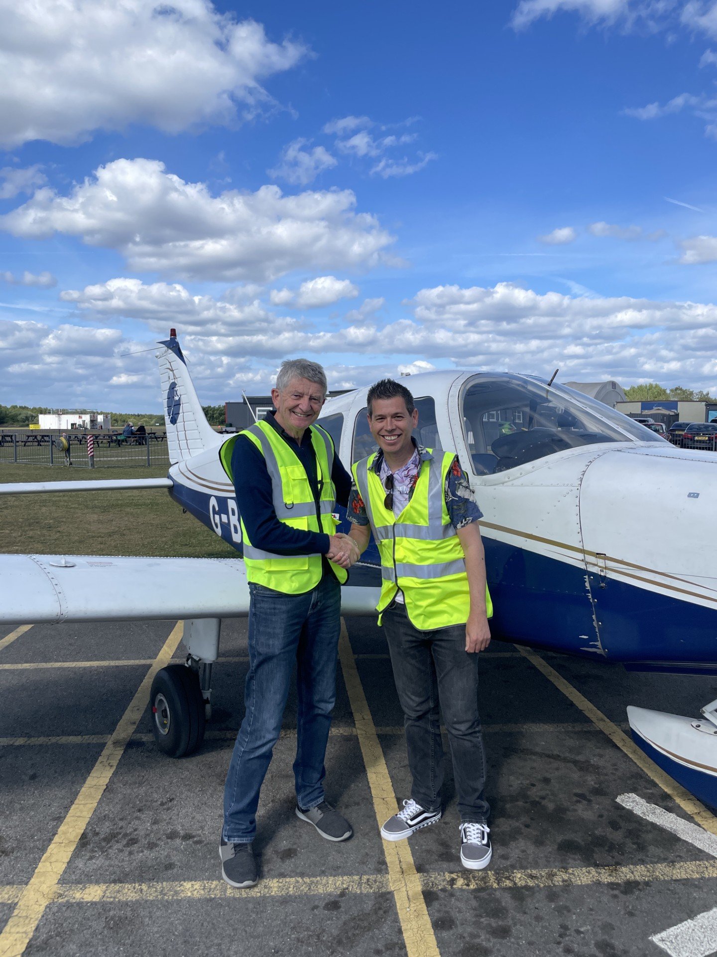 Two men wearing yellow reflective safety vests shaking hands in front of a small white and blue airplane on a tarmac, with a partly cloudy sky overhead.