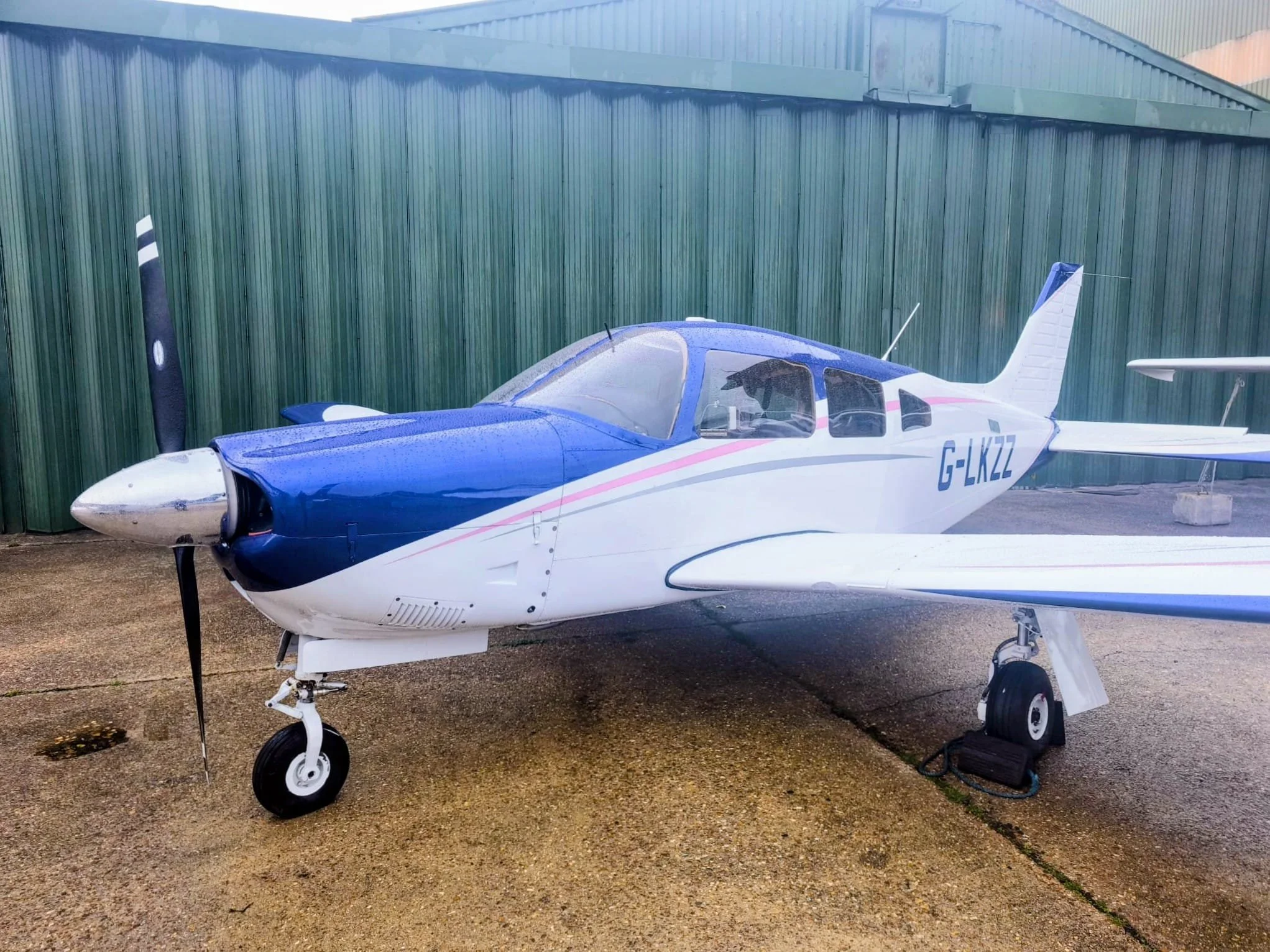 Small propeller aircraft with blue and white exterior, registration G-LKZZ, parked on tarmac in front of a green fence.