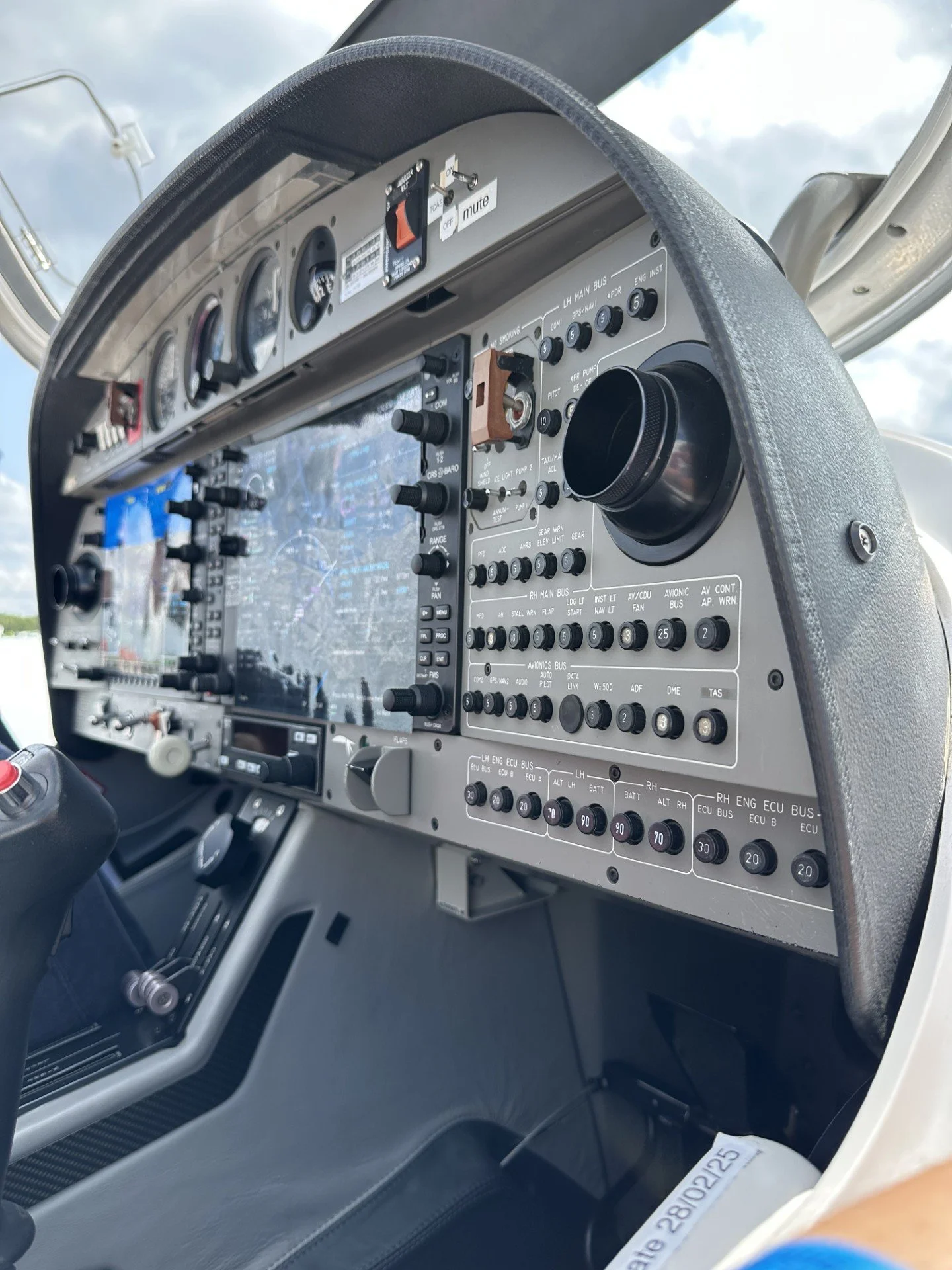 Cockpit dashboard of an aircraft with various gauges, switches, and controls, including a large screen in the center, set against a cloudy sky background.