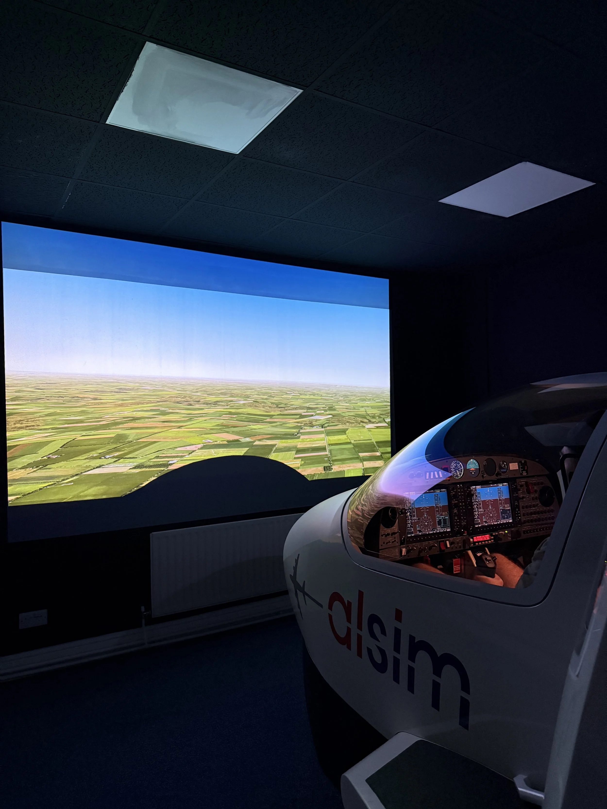 Flight simulator cockpit with a large screen displaying an aerial view of farmland, and a pilot seated inside the simulator.