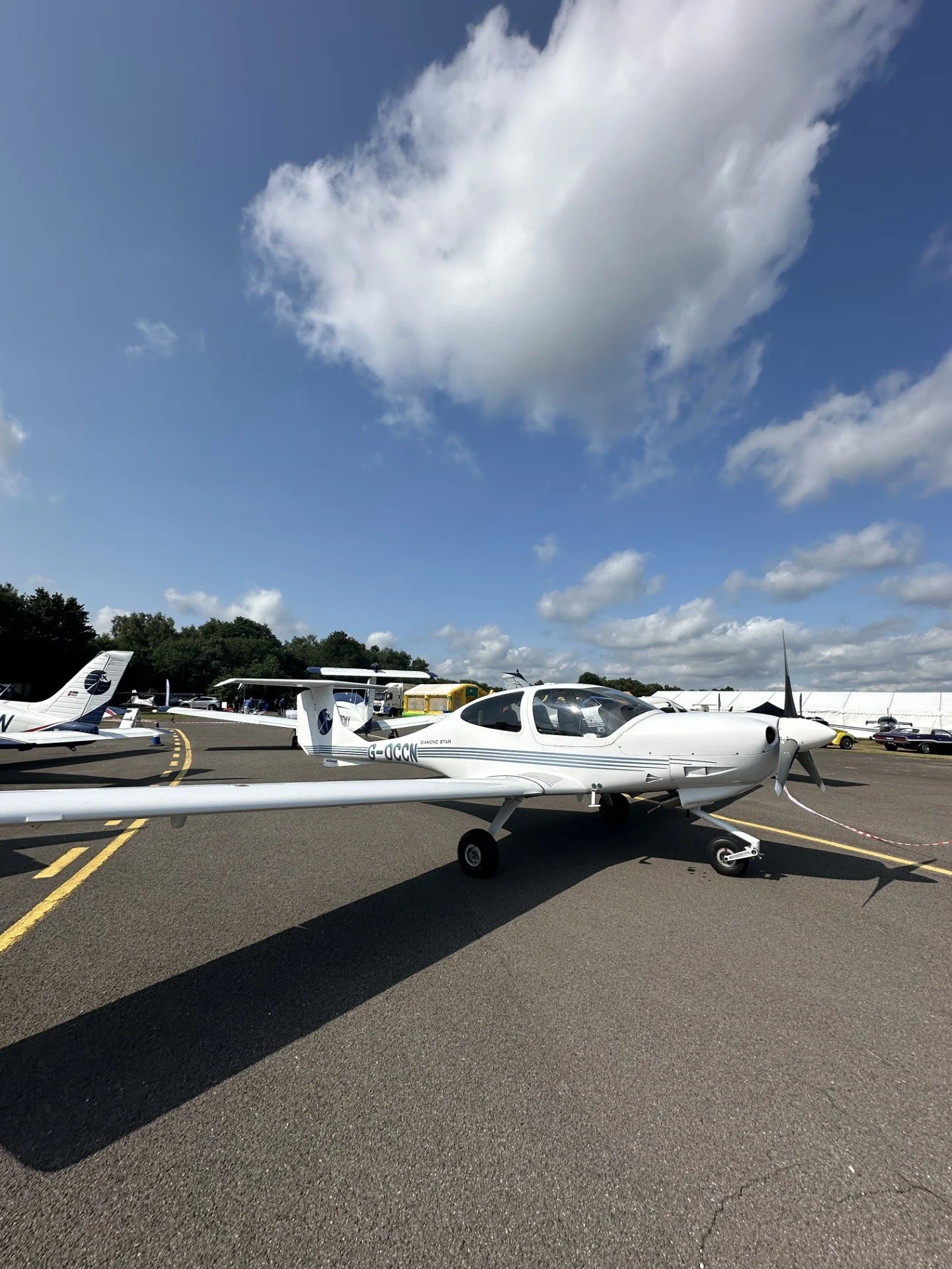 Small white aircraft on tarmac at an airfield with other planes and vehicles in the background, under a partly cloudy sky.