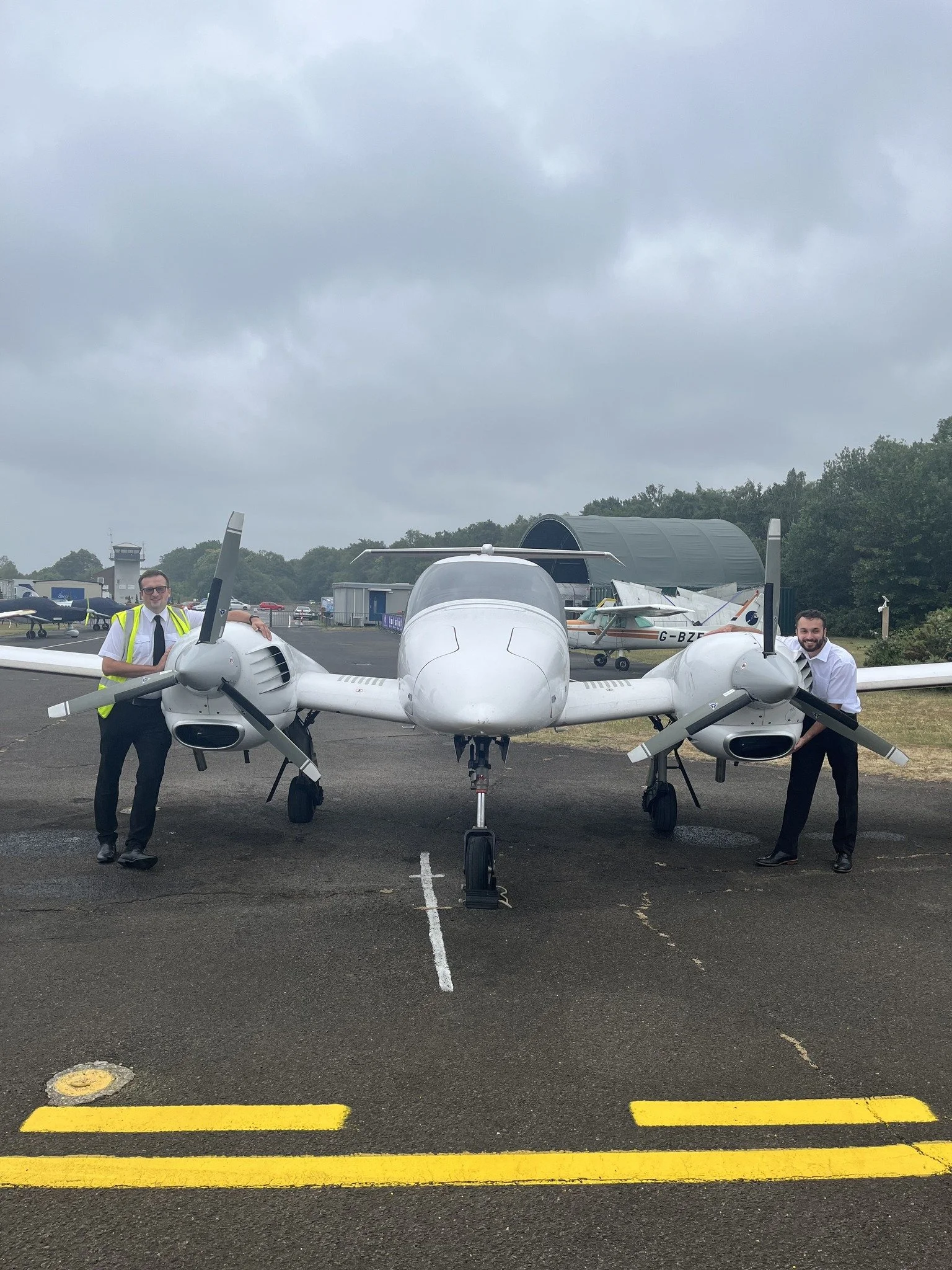 Two men standing next to a small white twin-engine aircraft on a tarmac, with planes and buildings in the background under cloudy sky.